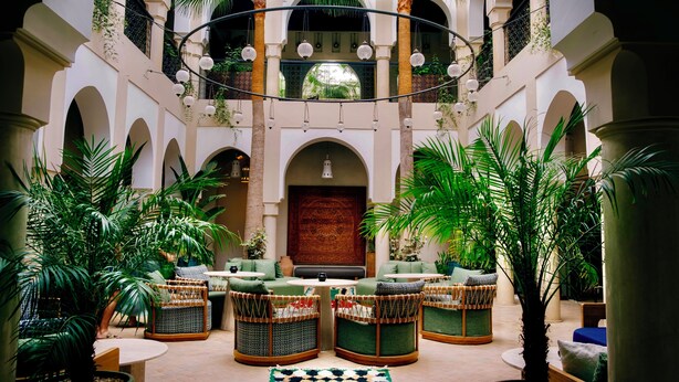 A spacious courtyard with multiple seating areas, a central wooden door, and a balcony with hanging lights. - Riad Nashira & Spa (Marrakech)