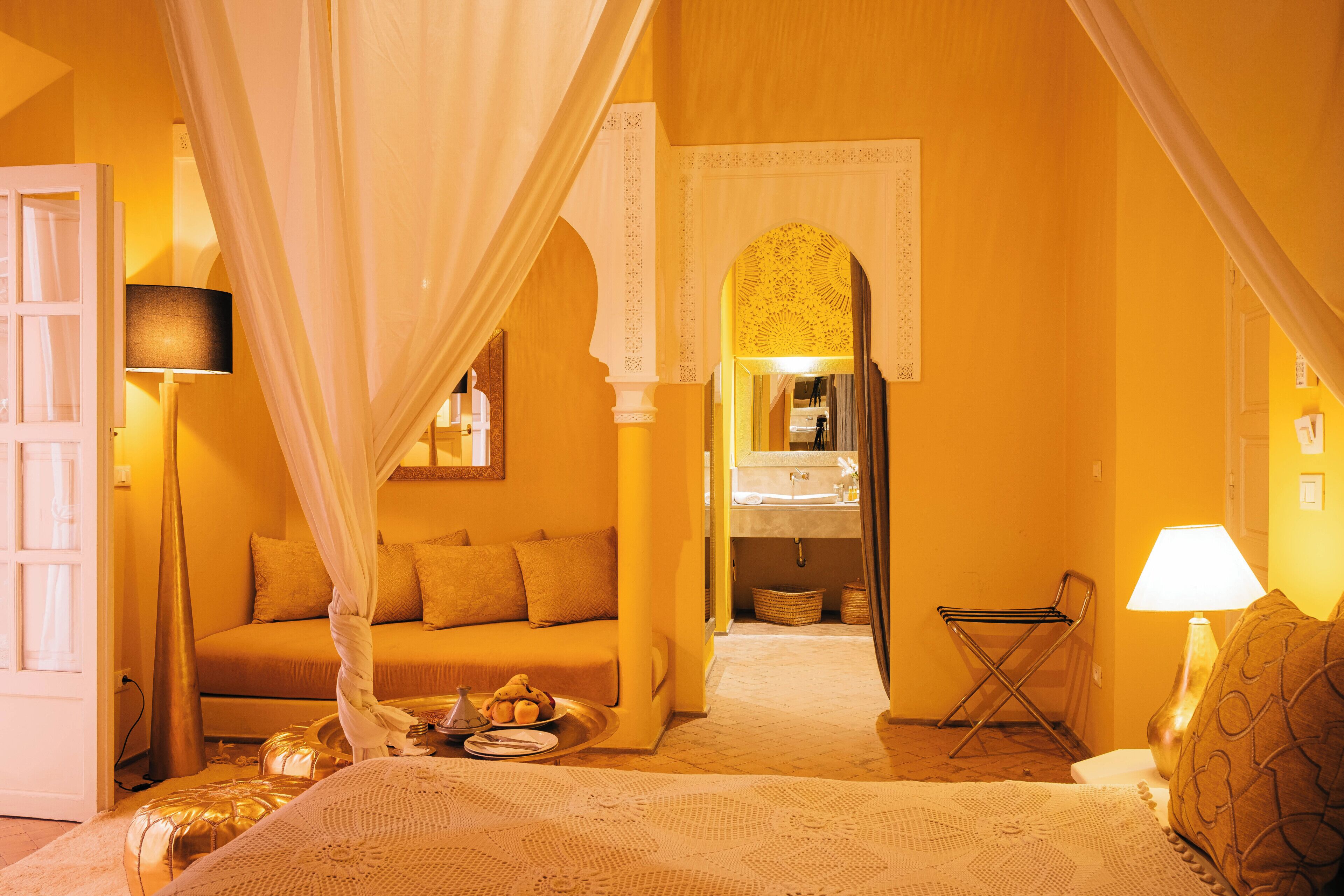 A room with a bed, a sofa, a bedside table with a lamp, a chair, and a small table with a bowl of fruit.