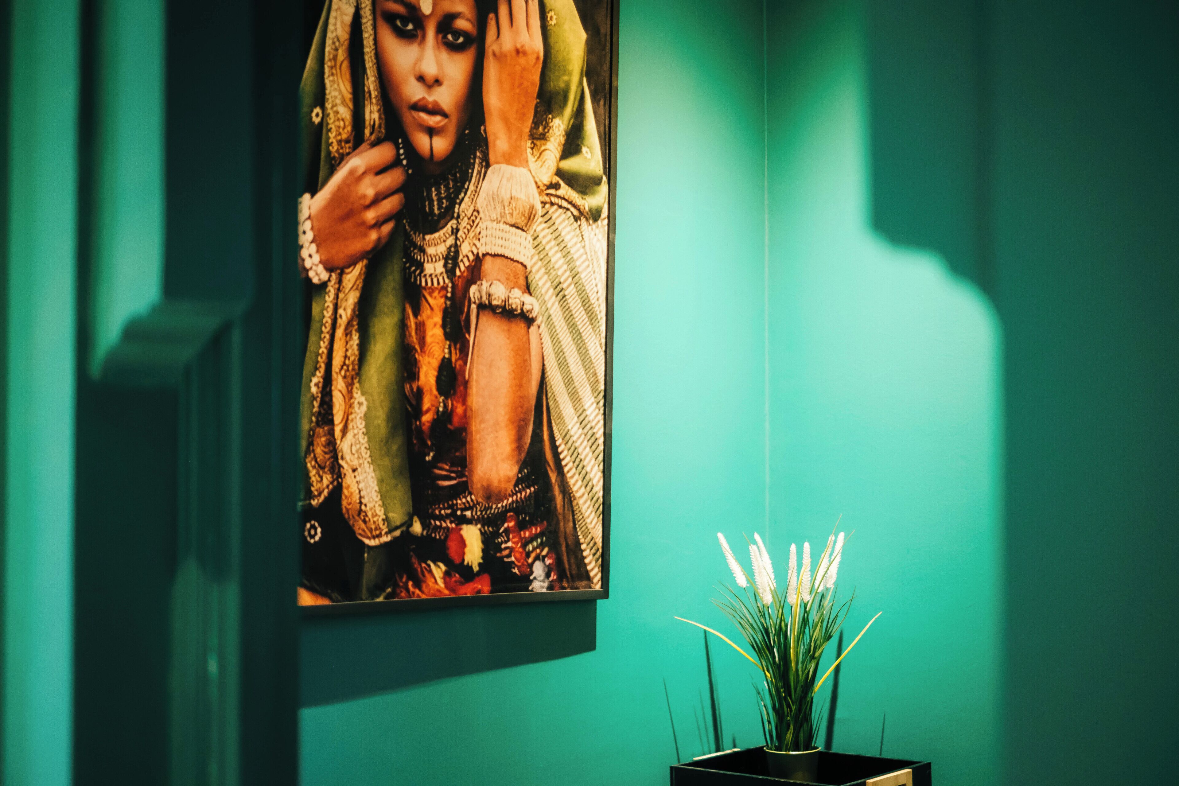 Une œuvre d’art encadrée sur un mur de couleur turquoise, un petit vase noir avec des fleurs blanches et un objet rectangulaire noir.