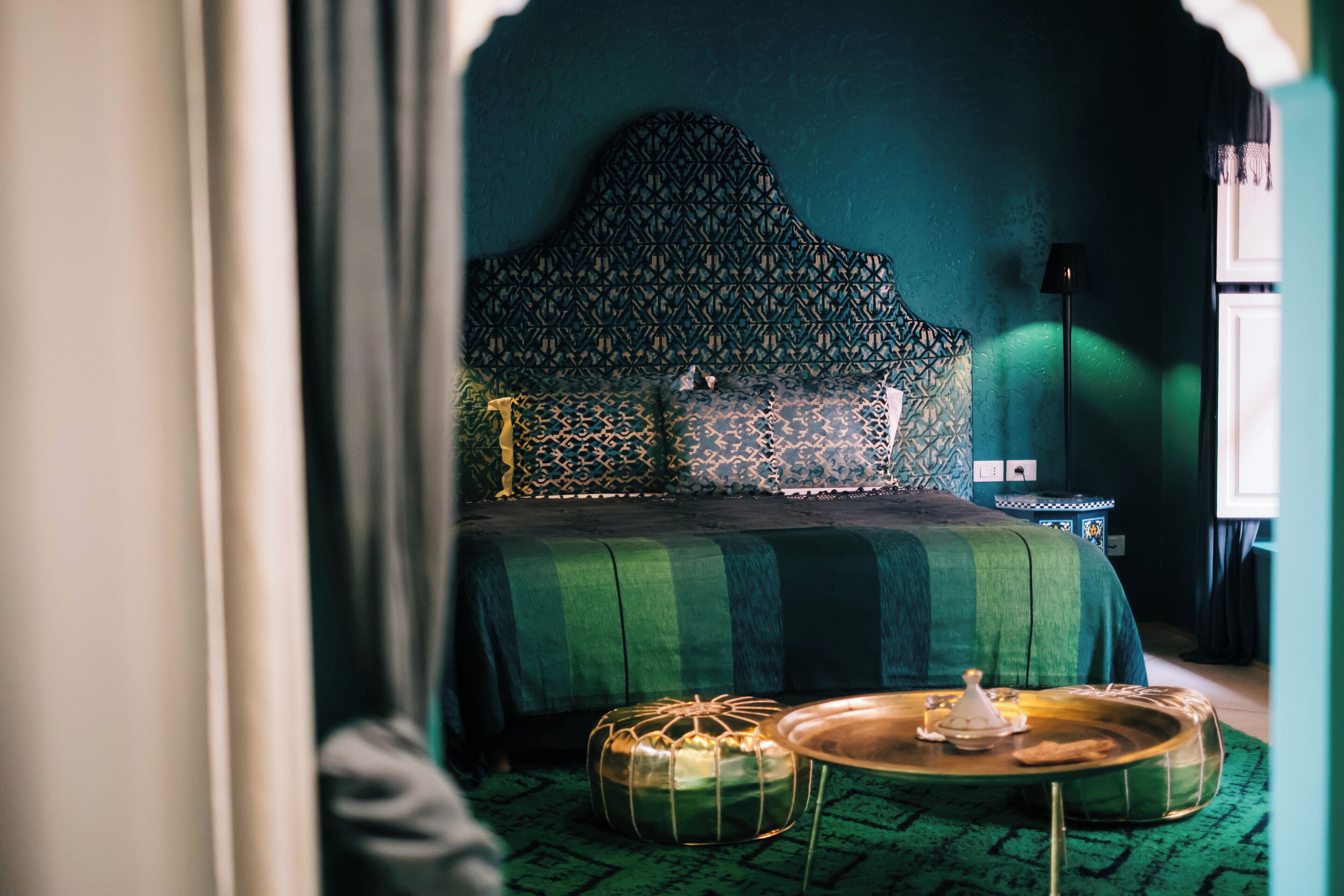 Una habitación con un cabecero estampado, una colcha verde, una mesa de centro redonda y un reposapiés estampado.