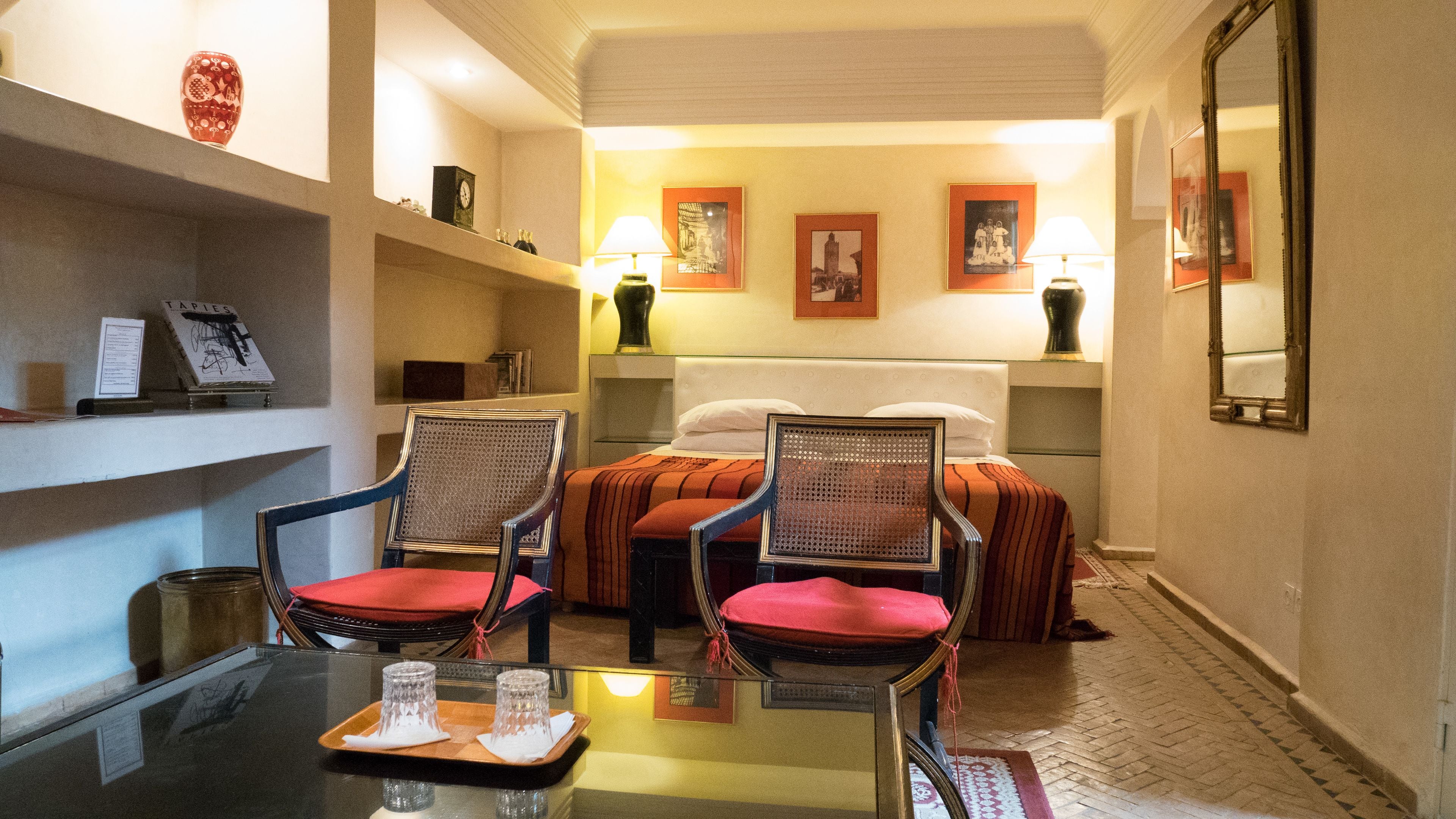 A hotel room with a bed, two chairs, a table with glasses, and framed pictures on the wall.
