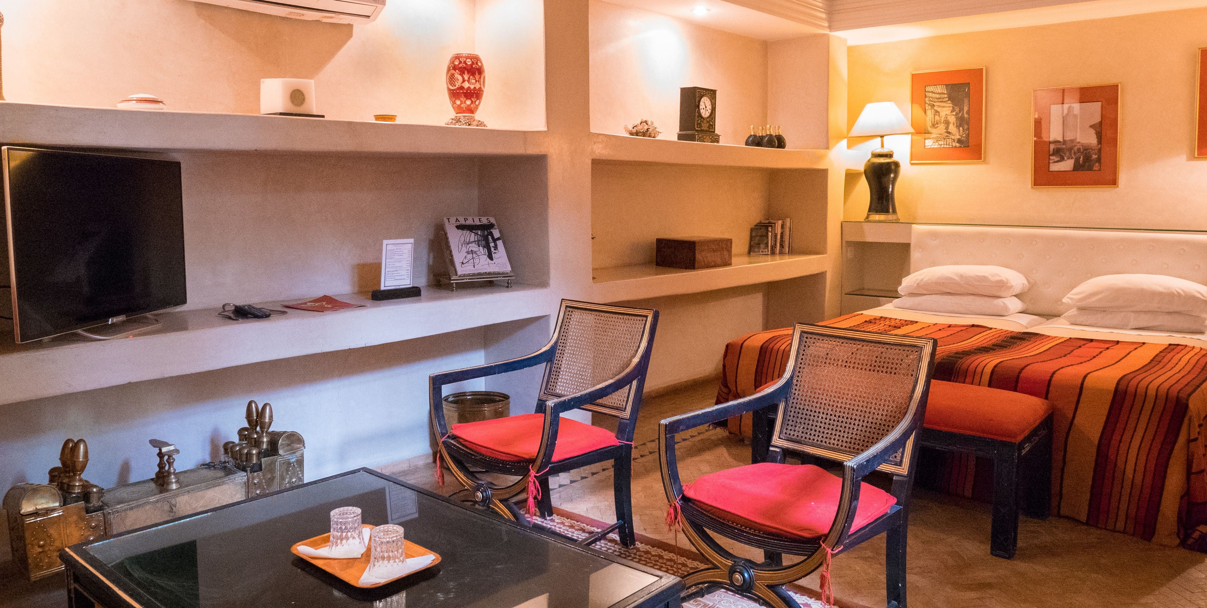 A hotel room with a bed, two chairs, a TV, and shelves with various items.