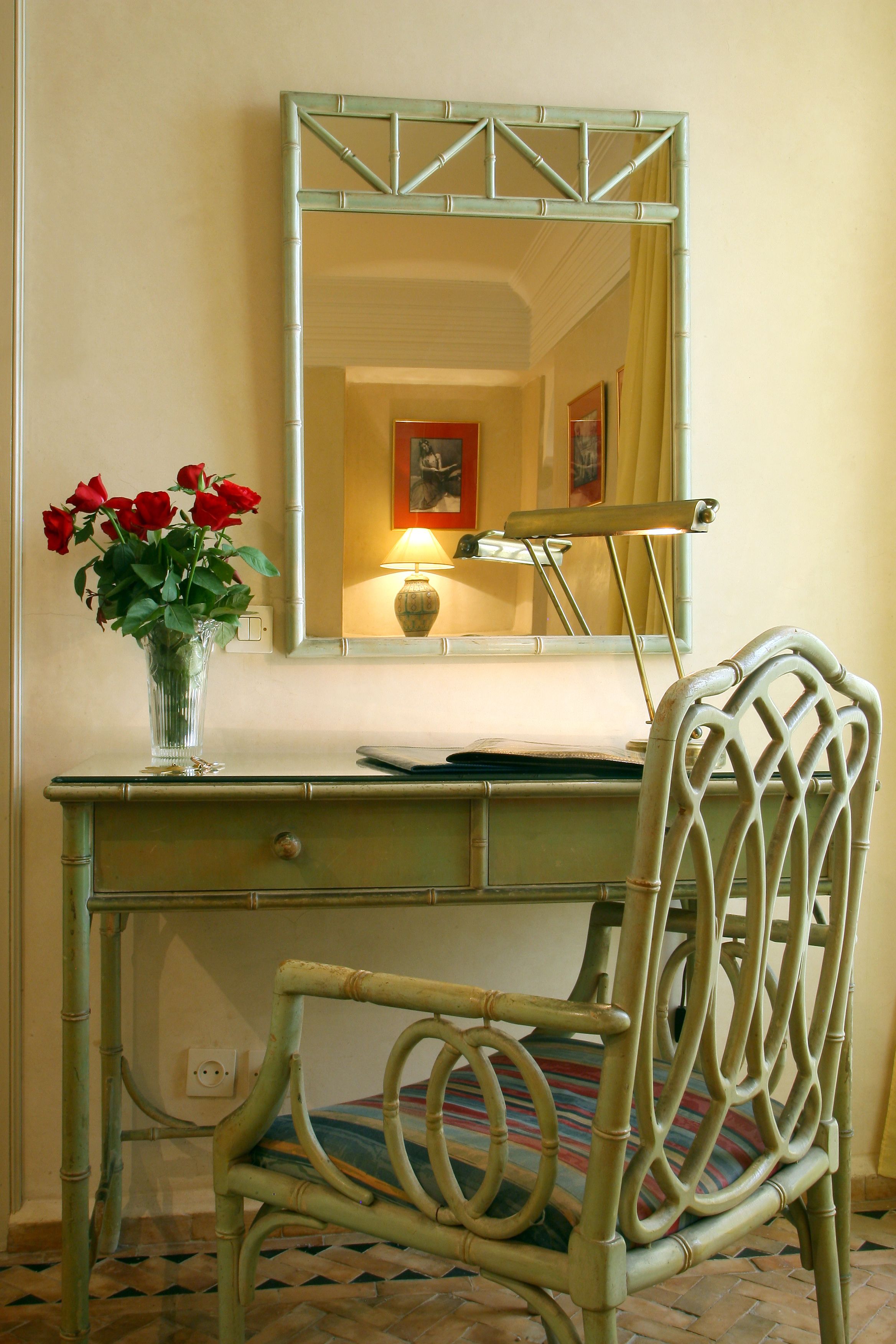 A room with a desk, chair, mirror, vase with flowers, lamp, and a bed with a colorful blanket.