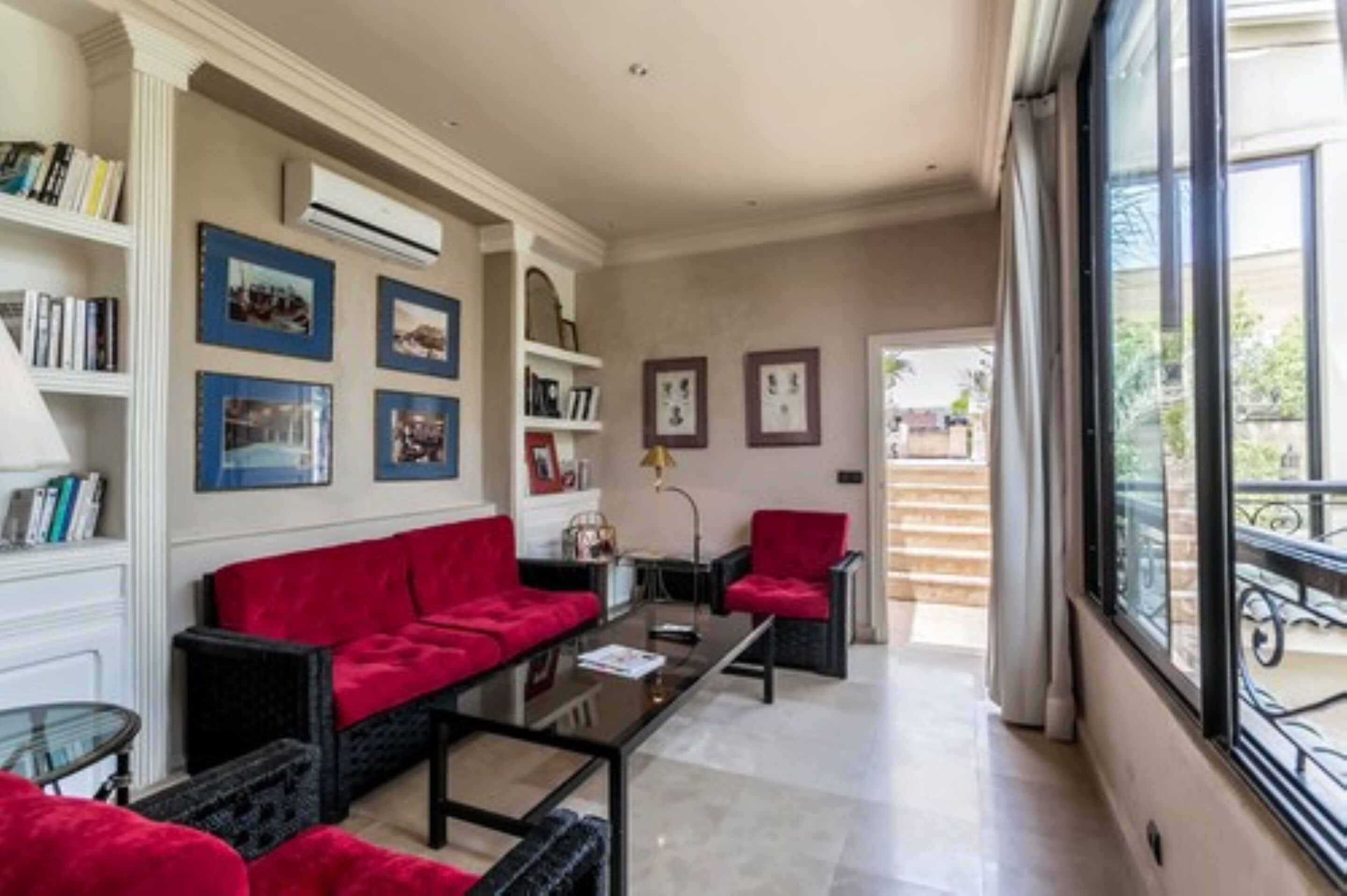 A spacious living room with a red sofa, black armchairs, and a glass coffee table. There are bookshelves with books and framed pictures on the walls.