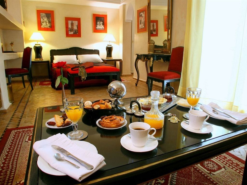 A hotel room with a dining table set for breakfast, a bed with red bedding, and a red armchair.