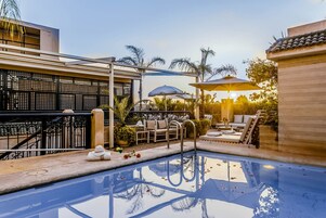 Una piscina con sillas de descanso y sombrillas, rodeada de palmeras y una pérgola.