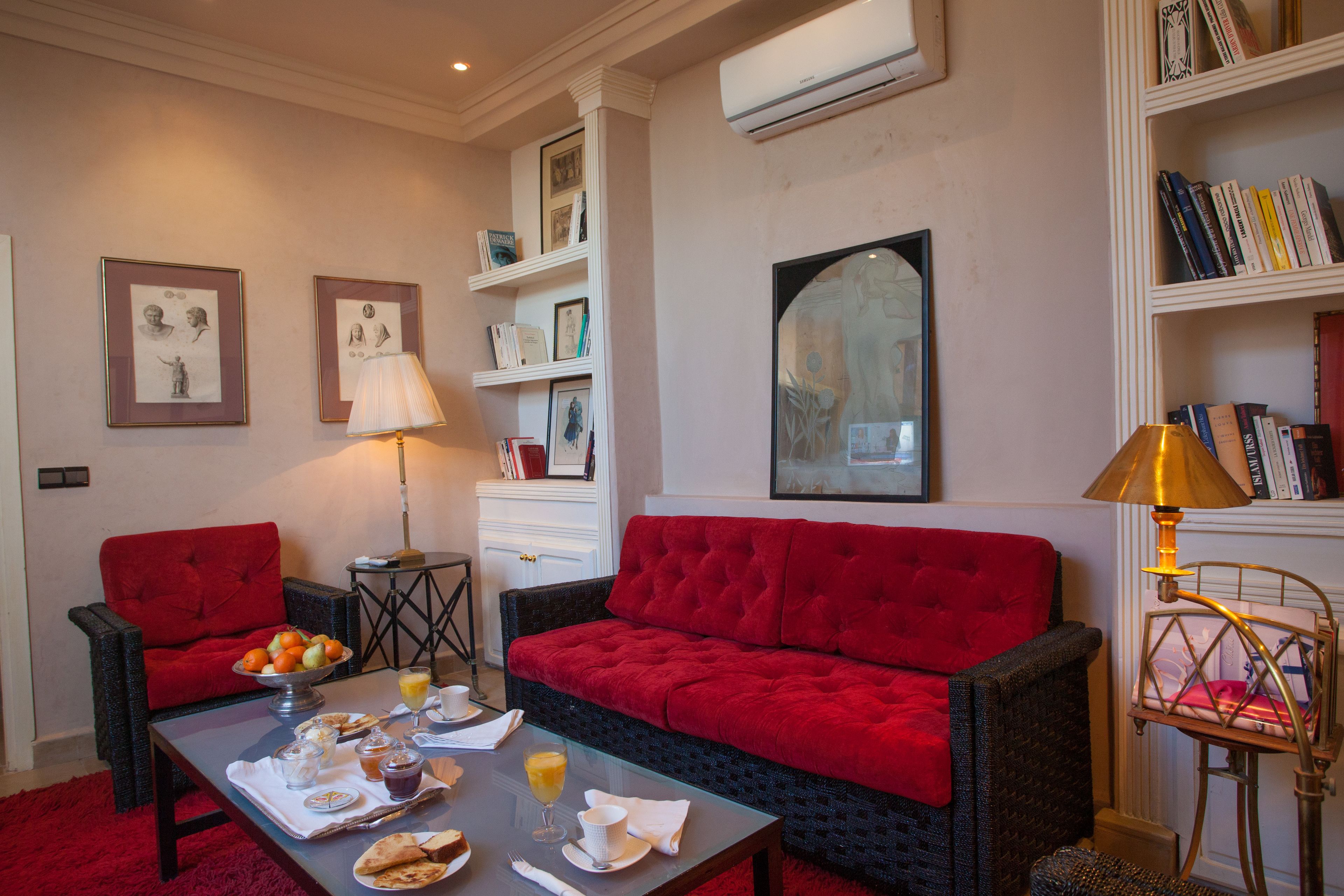 A living room with a red sofa, a coffee table with breakfast items, a bookshelf, and framed artwork on the walls.