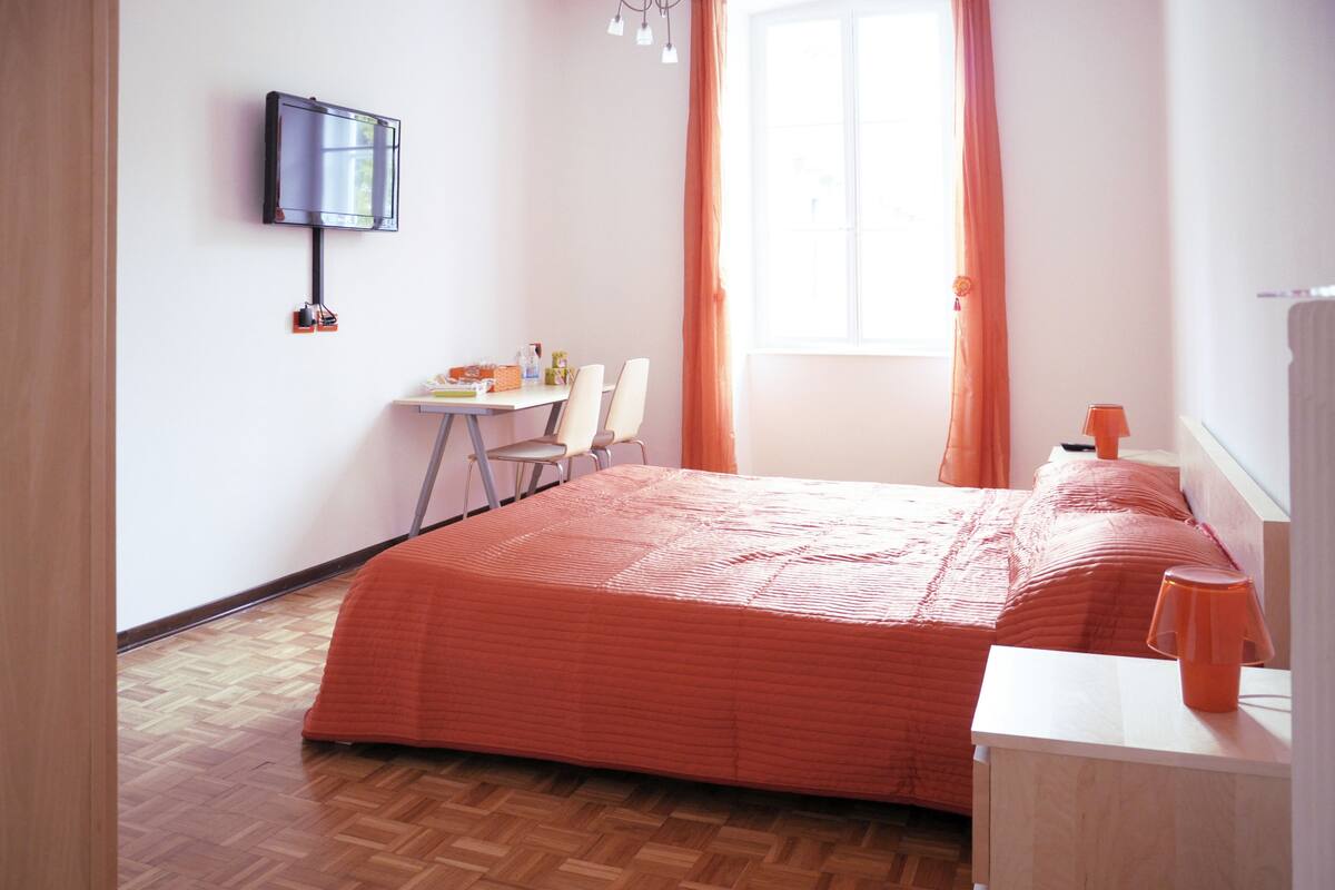 Quarto com cama, escrivaninha com cadeira, televisão na parede e janela com cortinas.