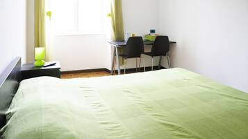 A neatly made bed with a green bedspread, a desk with two chairs, a lamp, and a window with curtains.
