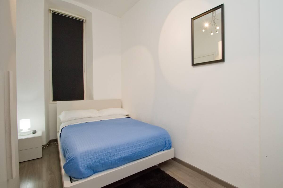 Um quarto com uma cama de solteiro, uma mesa de cabeceira, uma janela com persiana e um espelho fixado na parede.