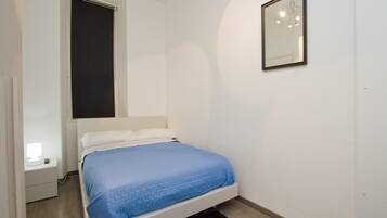 Une chambre avec un lit simple, une table de chevet, une fenêtre avec un store et un miroir fixé au mur.