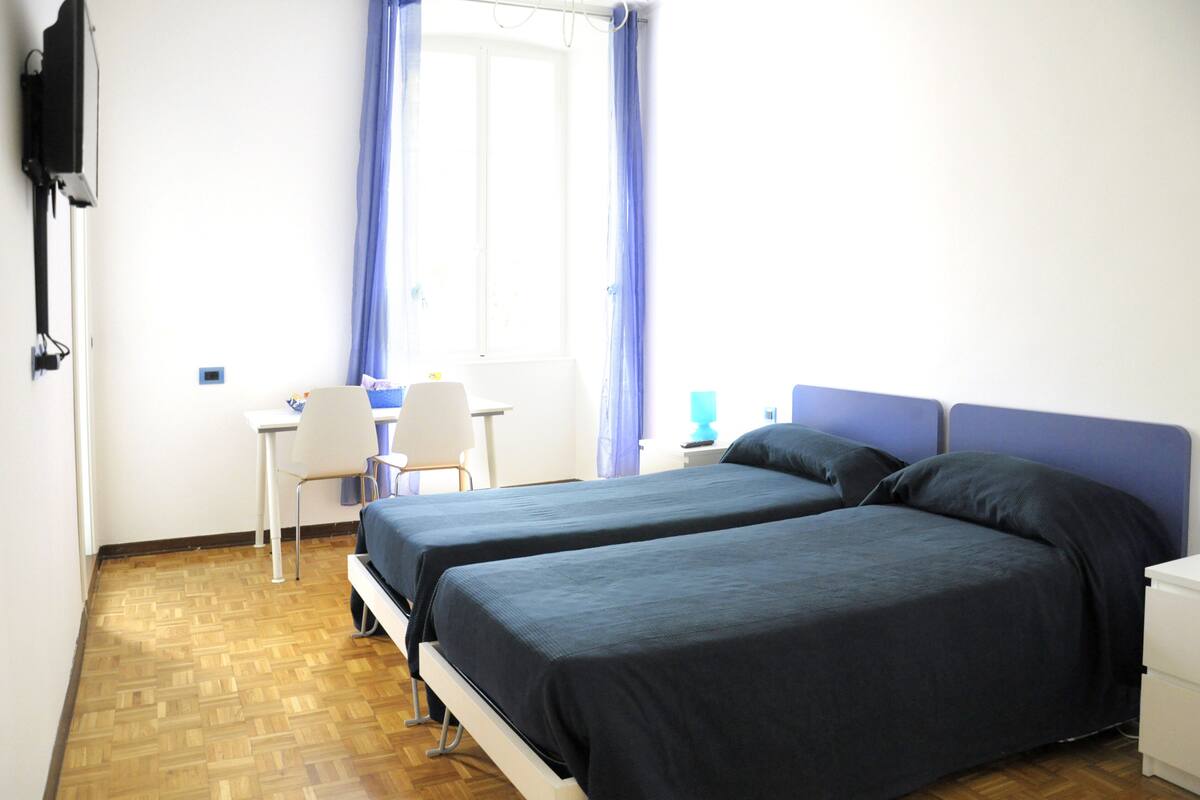 Quarto com duas camas, uma escrivaninha, uma cadeira e uma televisão.