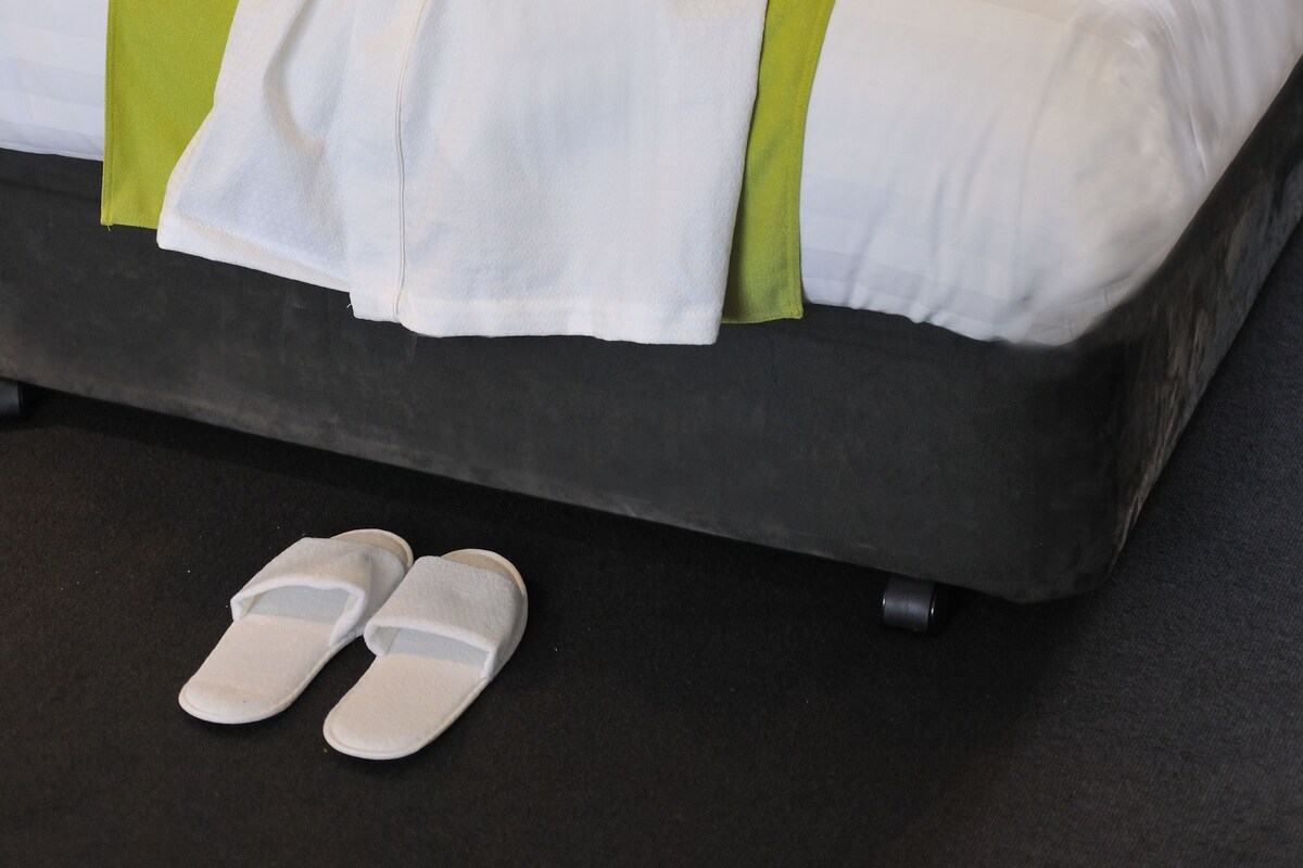 A neatly made bed with a green and white bedspread, a pair of slippers on the floor, and a chair with a green pillow in the background.