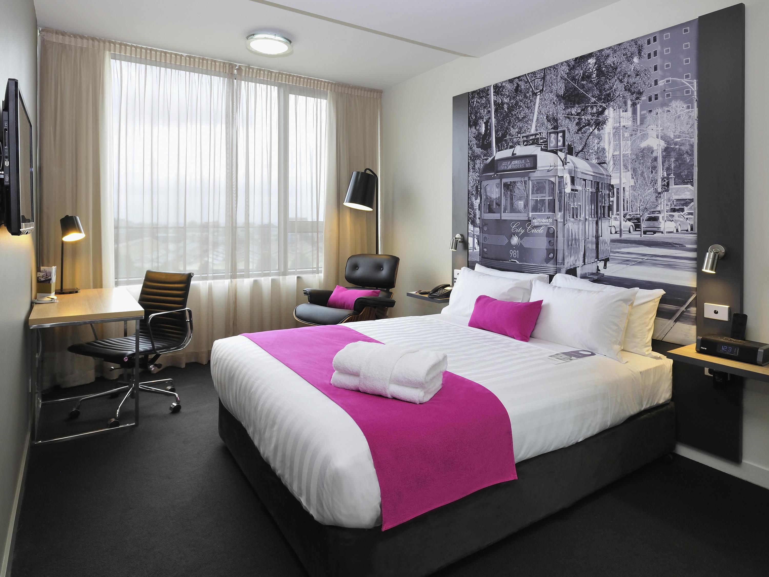 A hotel room with a large bed, a desk with a chair, a chair with a pillow, a desk lamp, and a wall-mounted TV.