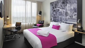 A hotel room with a large bed, a desk with a chair, a chair with a pillow, a desk lamp, and a wall-mounted TV.