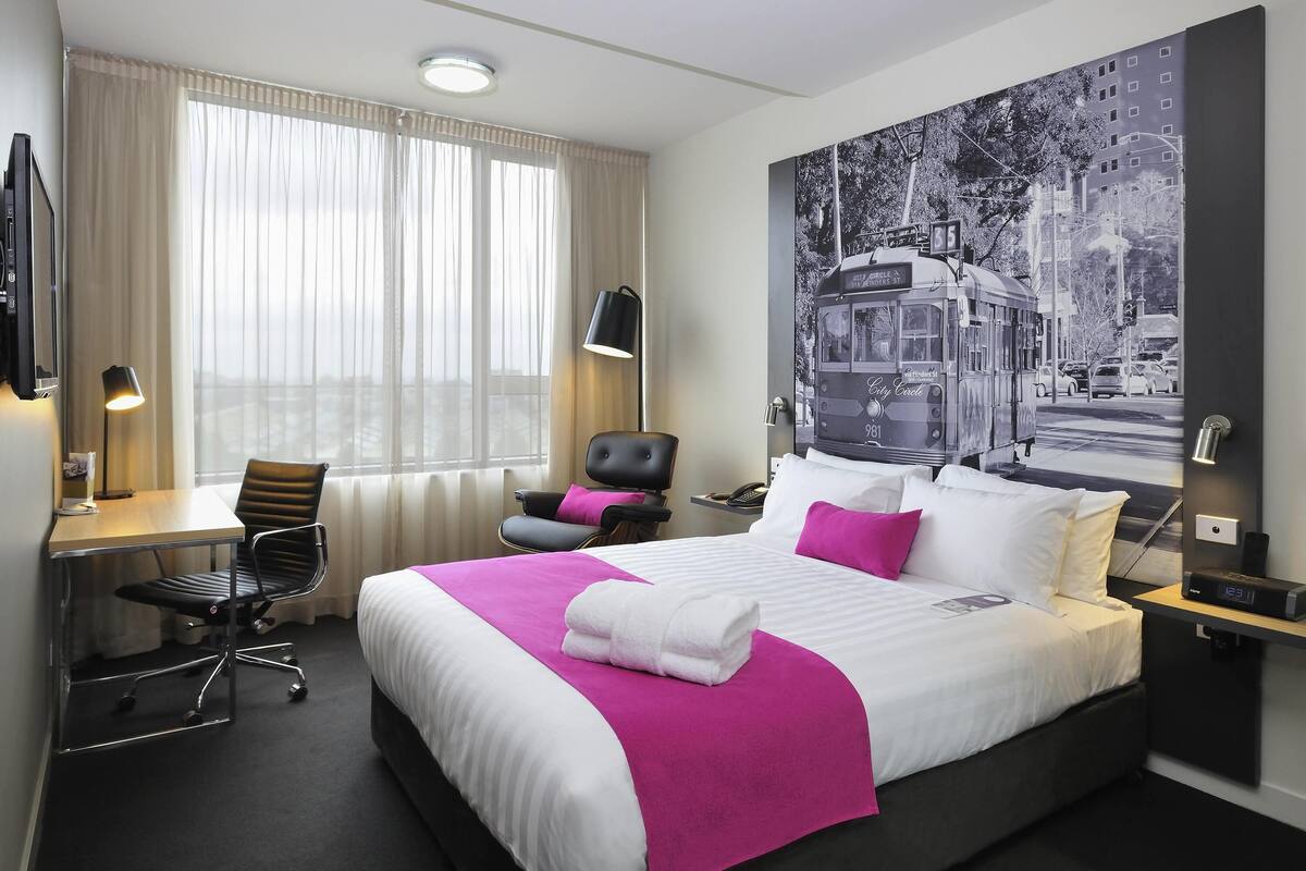 Ein Hotelzimmer mit einem großen Bett, einem Schreibtisch mit Stuhl, einem Sessel mit Kissen, einer Schreibtischlampe und einem an der Wand befestigten Fernseher.