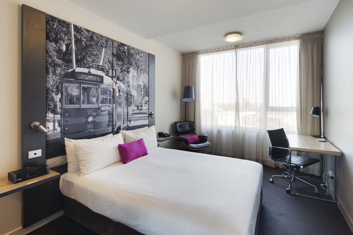 A hotel room with a large bed, a desk with a chair, a lamp, and a large poster on the wall.