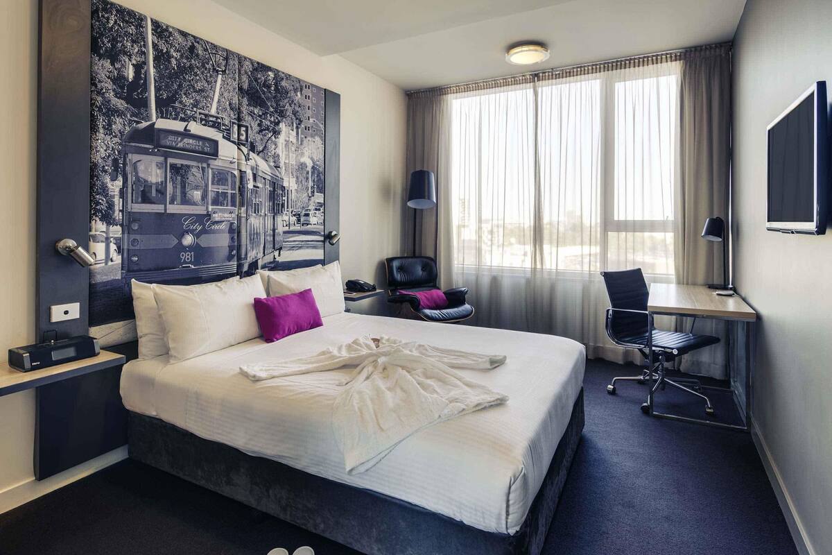Ein Hotelzimmer mit Bett, Schreibtisch, Stuhl, Fernseher und einem großen Bild an der Wand.