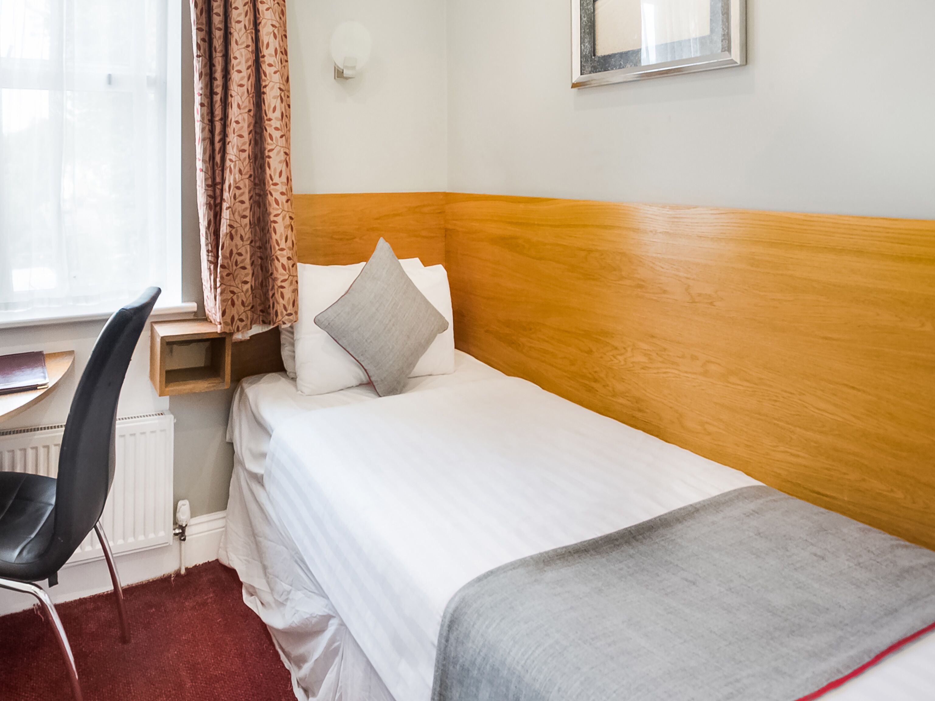 A hotel room with a single bed, a wooden headboard, a desk with a chair, a radiator, and a window with curtains.