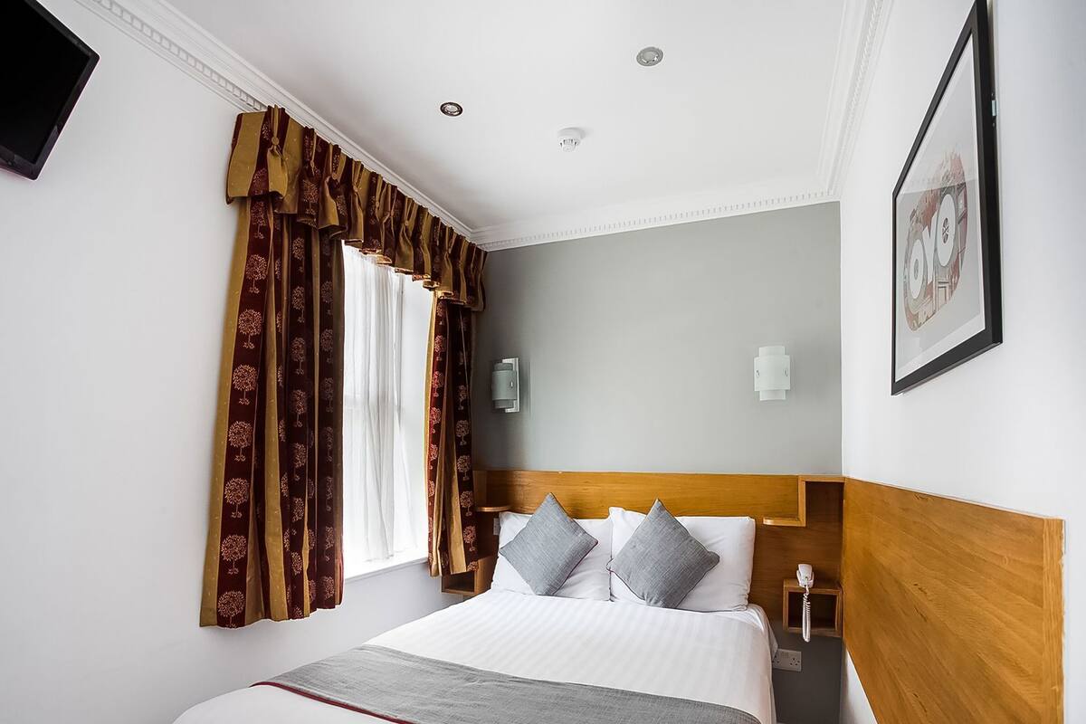 A hotel room with a bed, curtains, a headboard, a bedside table, a wall-mounted light, and a framed picture.
