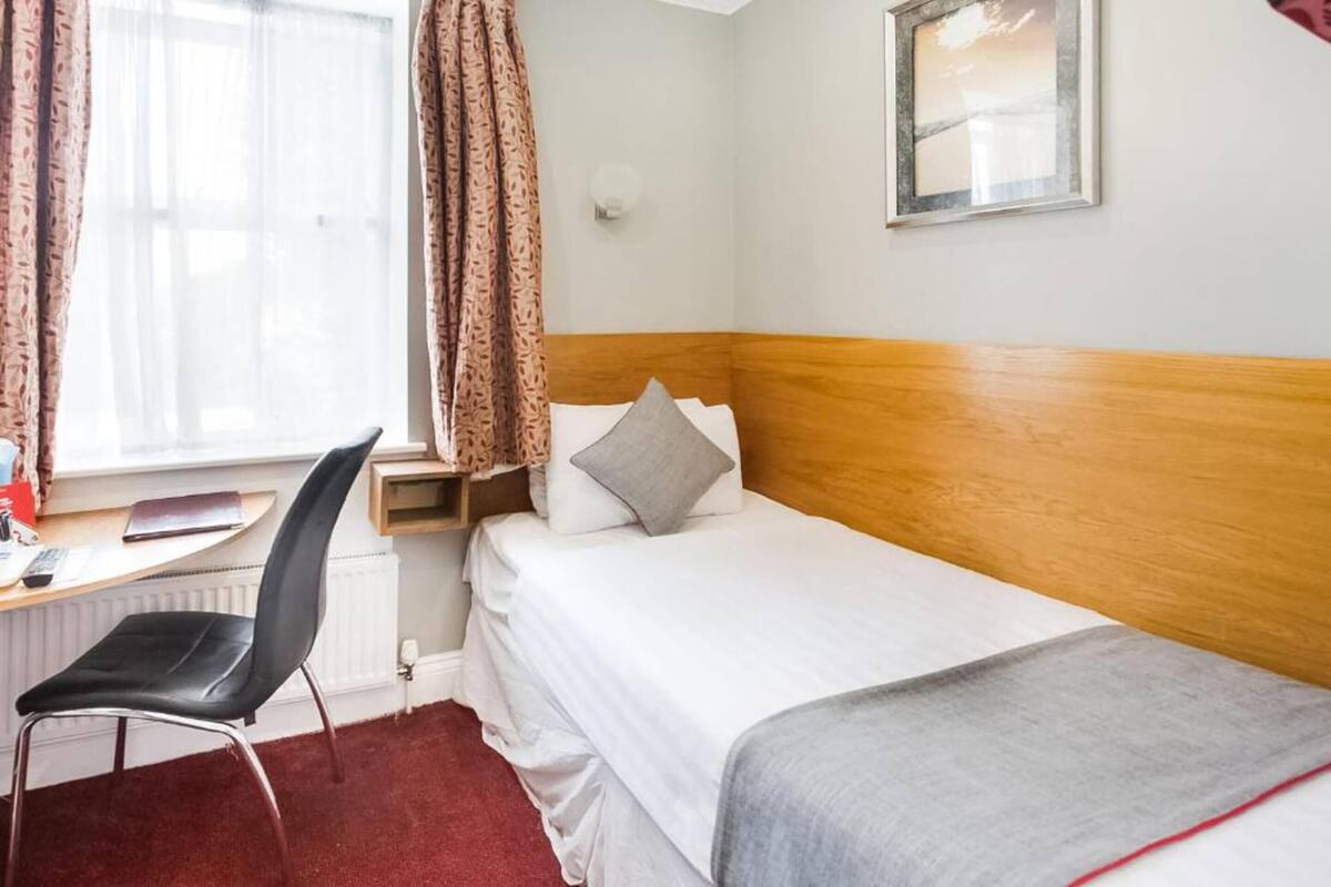 A hotel room with a bed, a desk with a chair, a window with curtains, and a wall-mounted light.