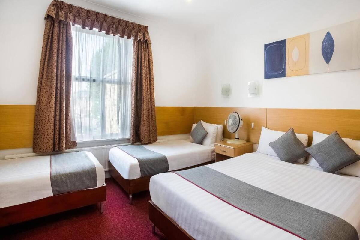 A hotel room with two single beds, a large bed, a window with curtains, a bedside table with a lamp, and a wall art piece.