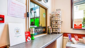 A hotel reception area with a reception desk, a clock, a TripAdvisor certificate, a map stand, and a refrigerator with beverages. - London Guest House by OYO - Acton (London)