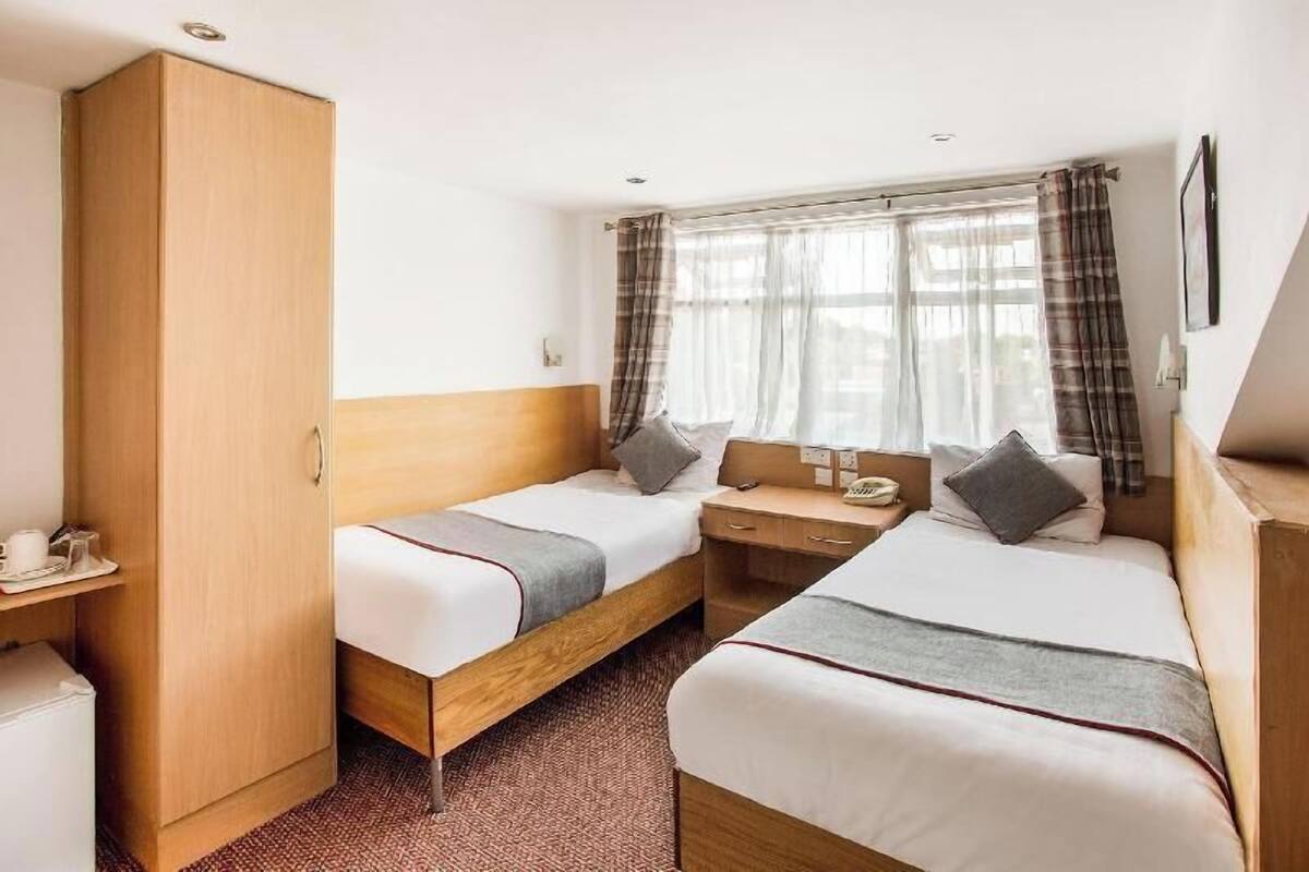 A hotel room with two single beds, a wooden wardrobe, a desk with a telephone, and a window with curtains.