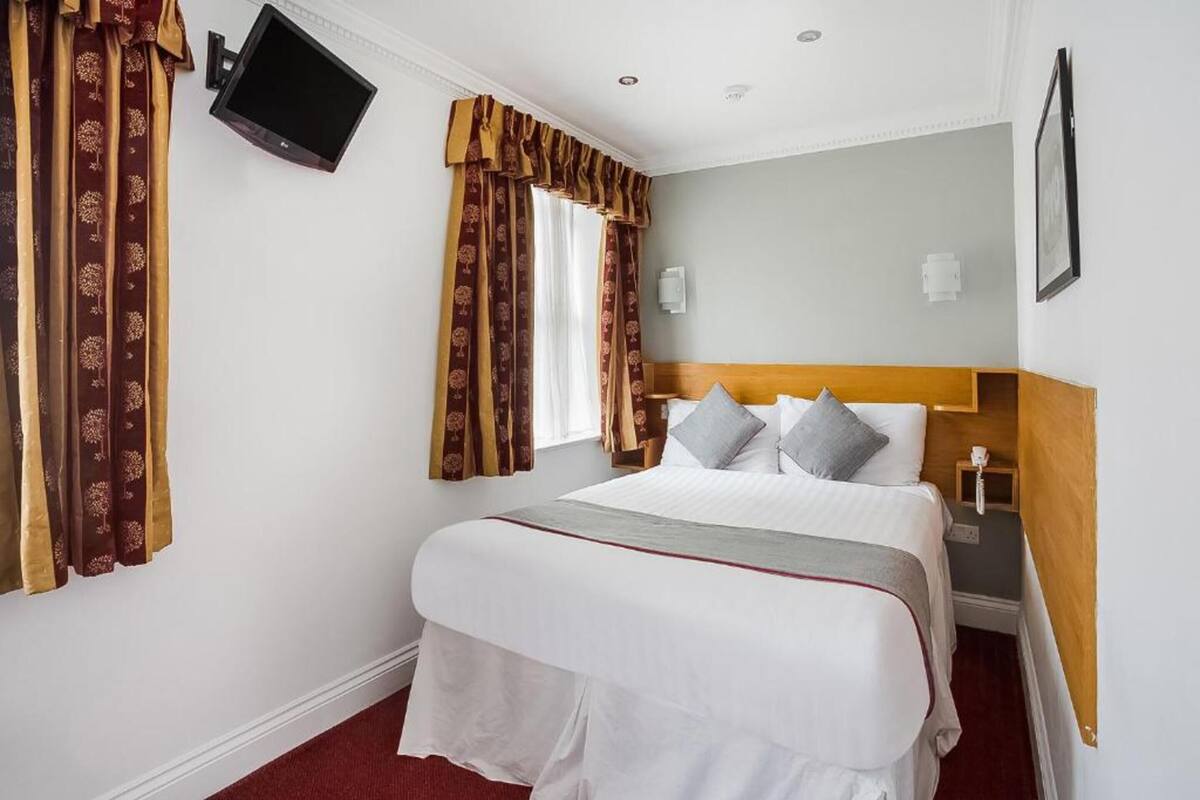A hotel room with a bed, curtains, a TV, a bedside table, and a wall-mounted light.