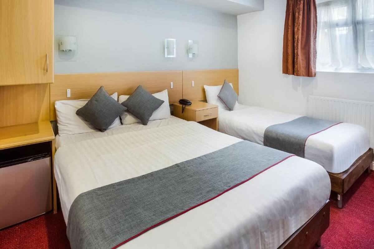 A hotel room with two beds, a wooden wardrobe, a bedside table with a lamp, and a window with curtains.