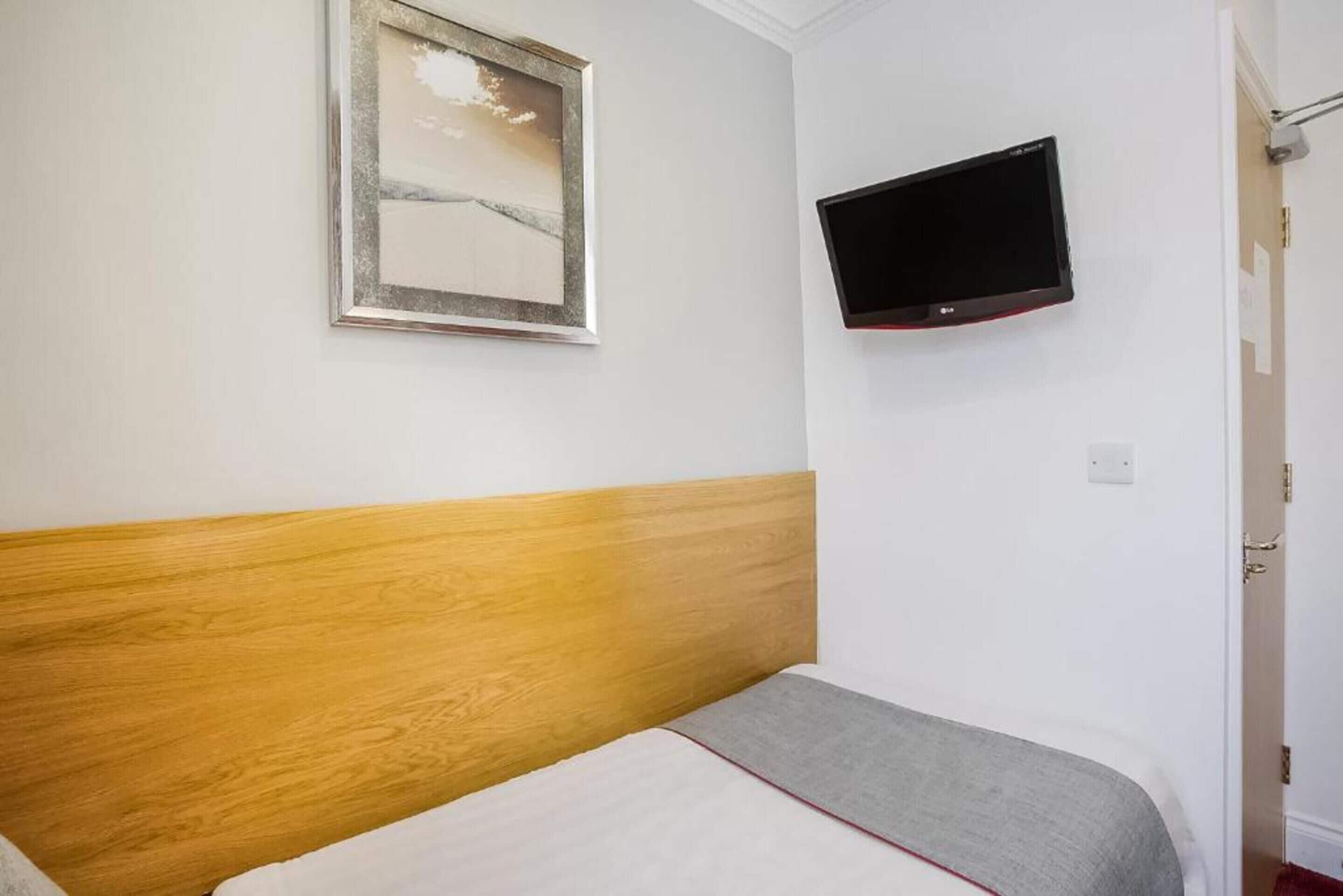 A room with a wooden headboard, a bed with a grey and white bedspread, a framed picture on the wall, and a mounted TV.