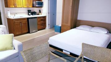 A compact hotel room with a bed, a small kitchenette, a seating area with a chair, and a wooden wardrobe.