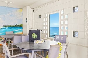 Ein Zimmer mit einem runden Tisch, weißen Stühlen, einem Bild mit Strandmotiv, einem Fenster mit Meerblick und einer Tasche mit dem Aufdruck 'Imperial'.