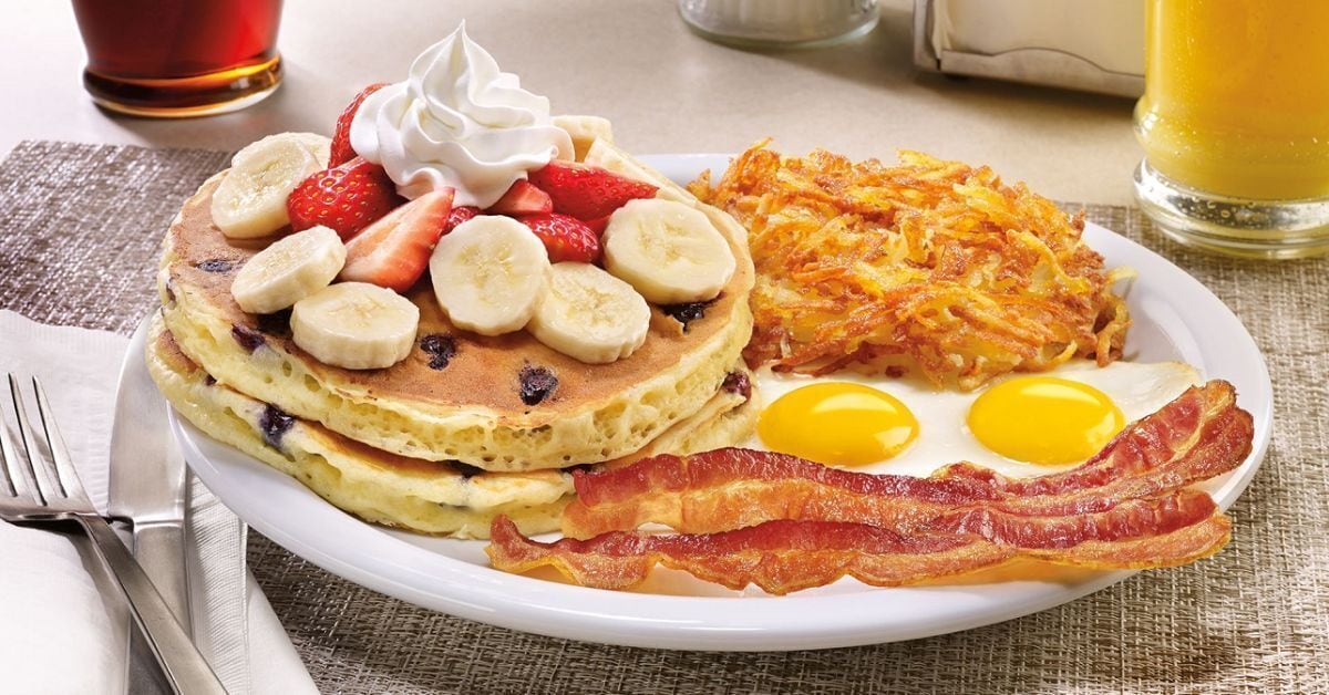 A breakfast plate with pancakes, strawberries, bananas, blueberries, hash browns, eggs, and bacon.