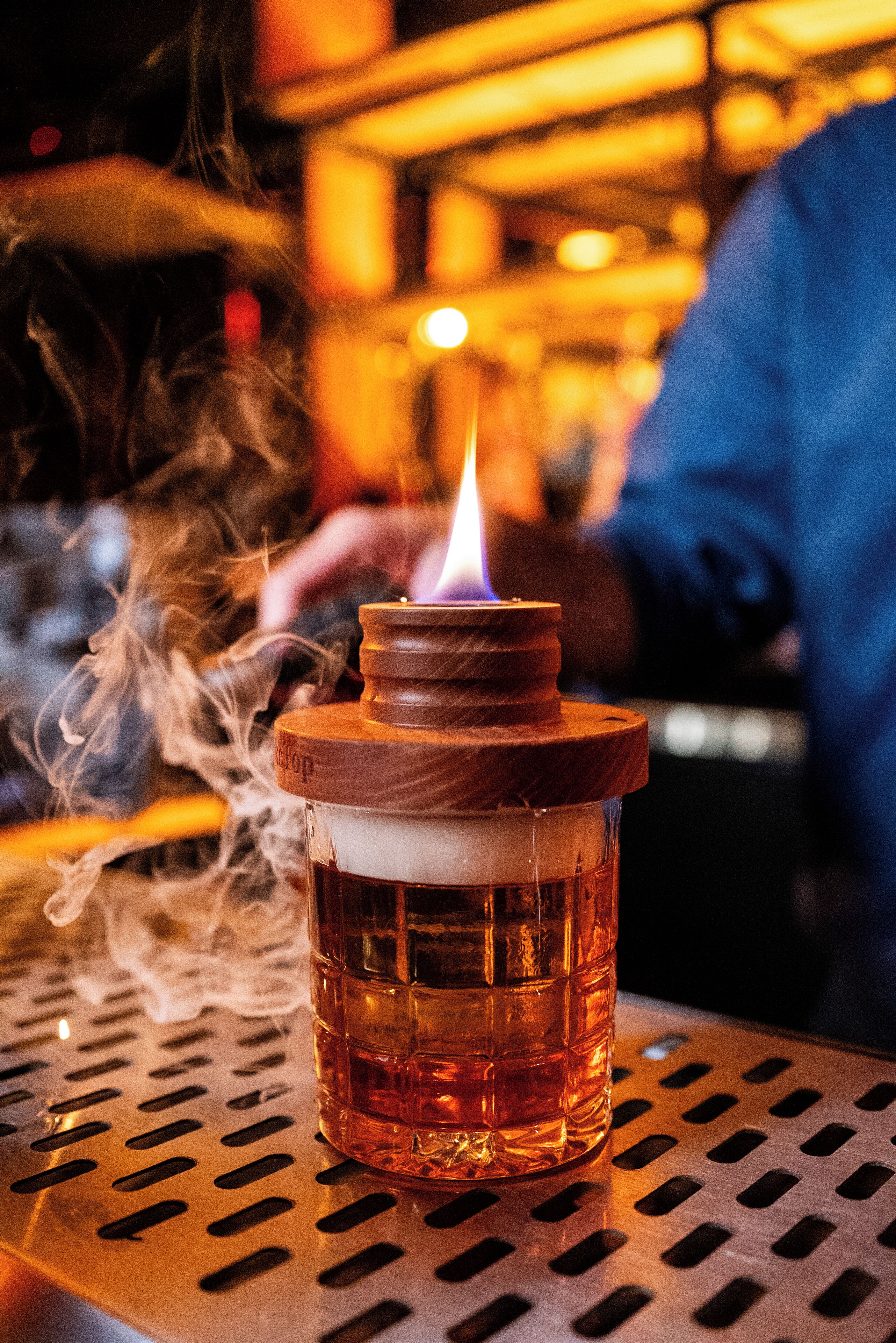 A close-up of a glass bottle with a wooden lid and a lit candle inside, placed on a perforated metal surface.