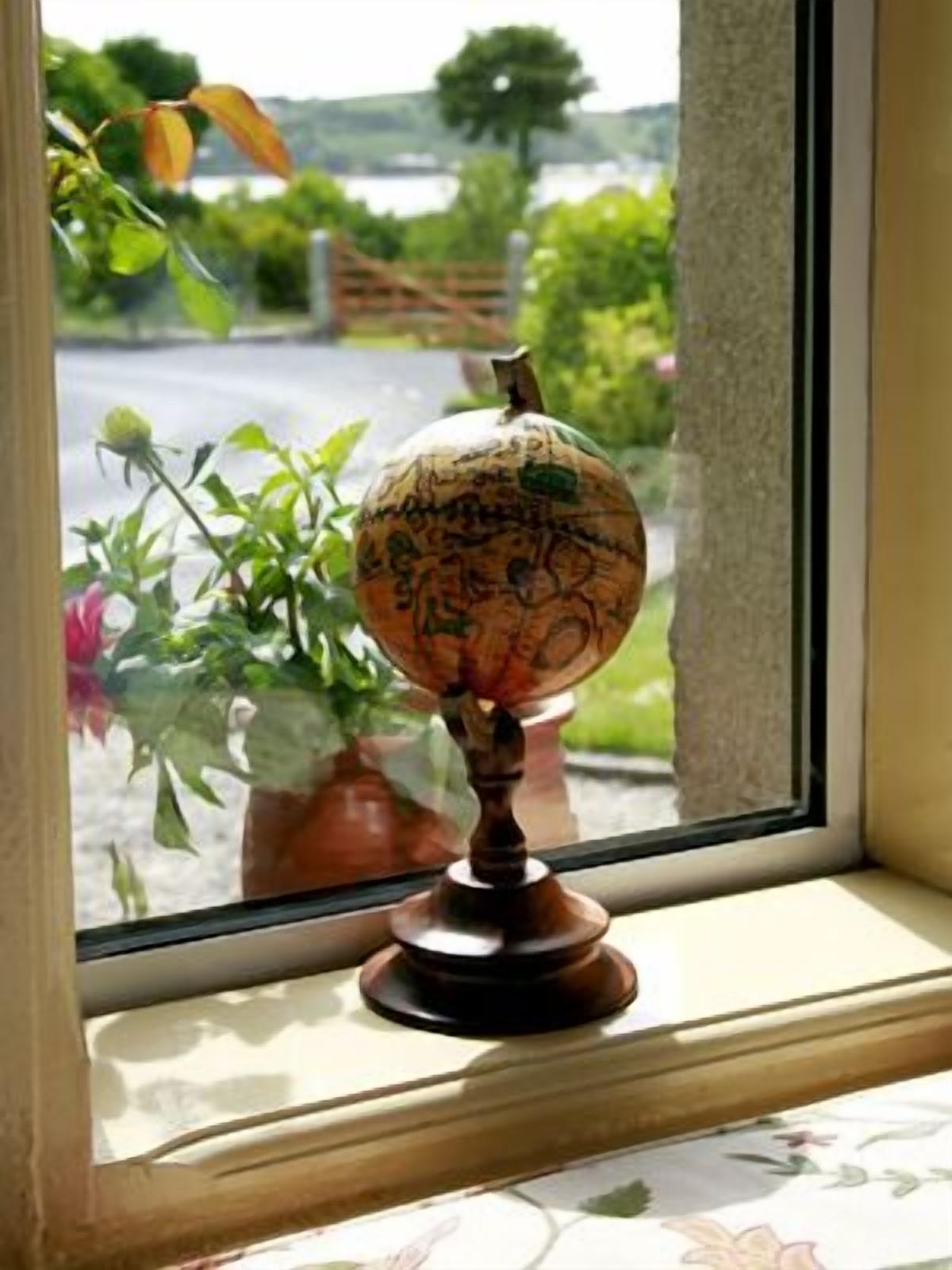 A globe on a wooden stand by a window with a view of greenery and a road.