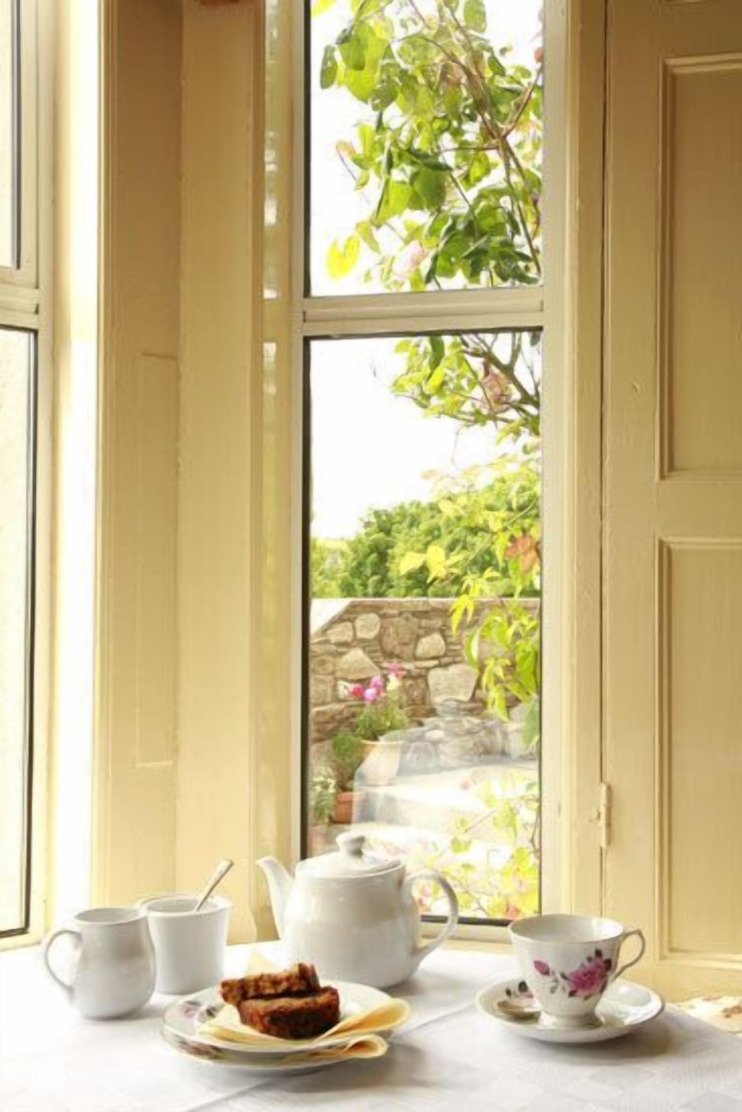 A table set with a teapot, cups, saucer, and a plate with a pastry, positioned by a window with a view of greenery and a stone wall.