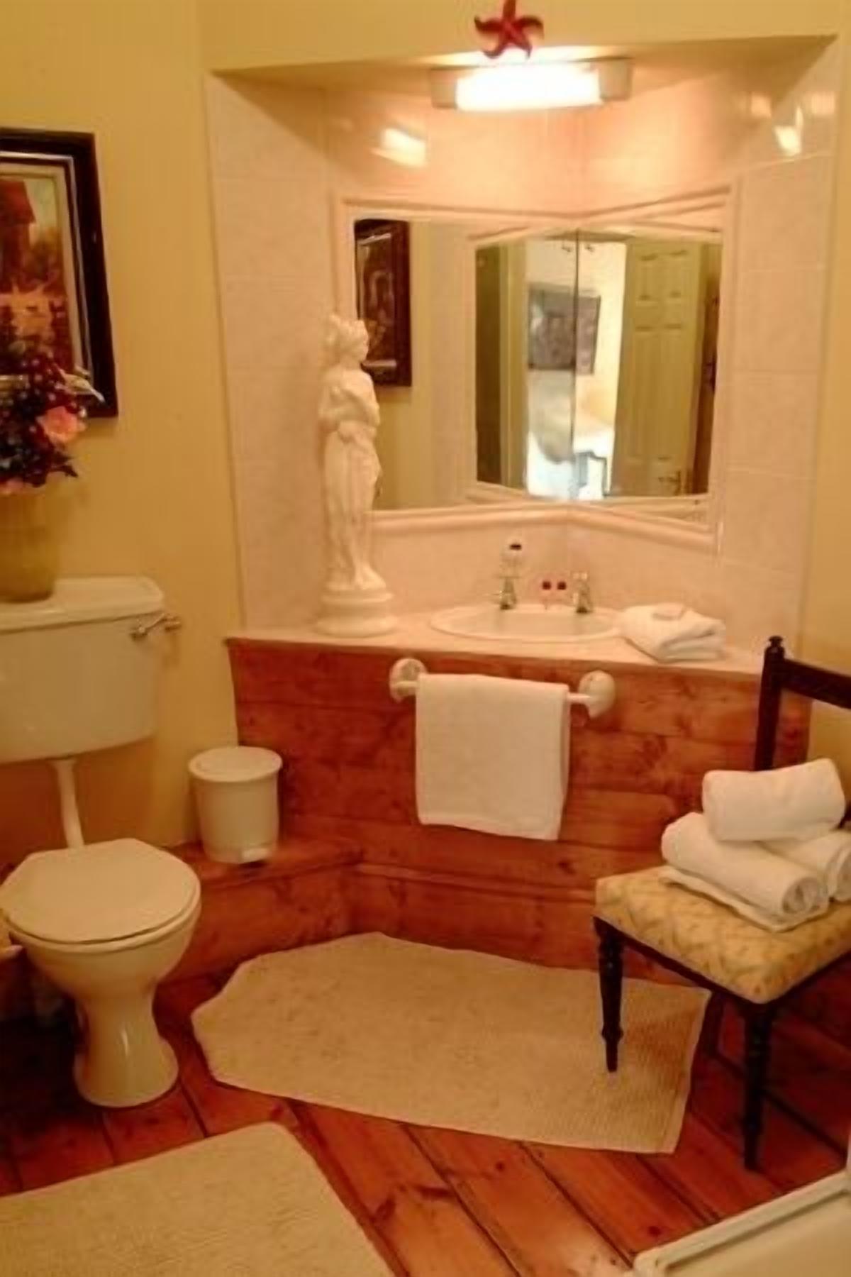 Un baño con tocador de madera, inodoro blanco, lavamanos con espejo y una silla con toallas.