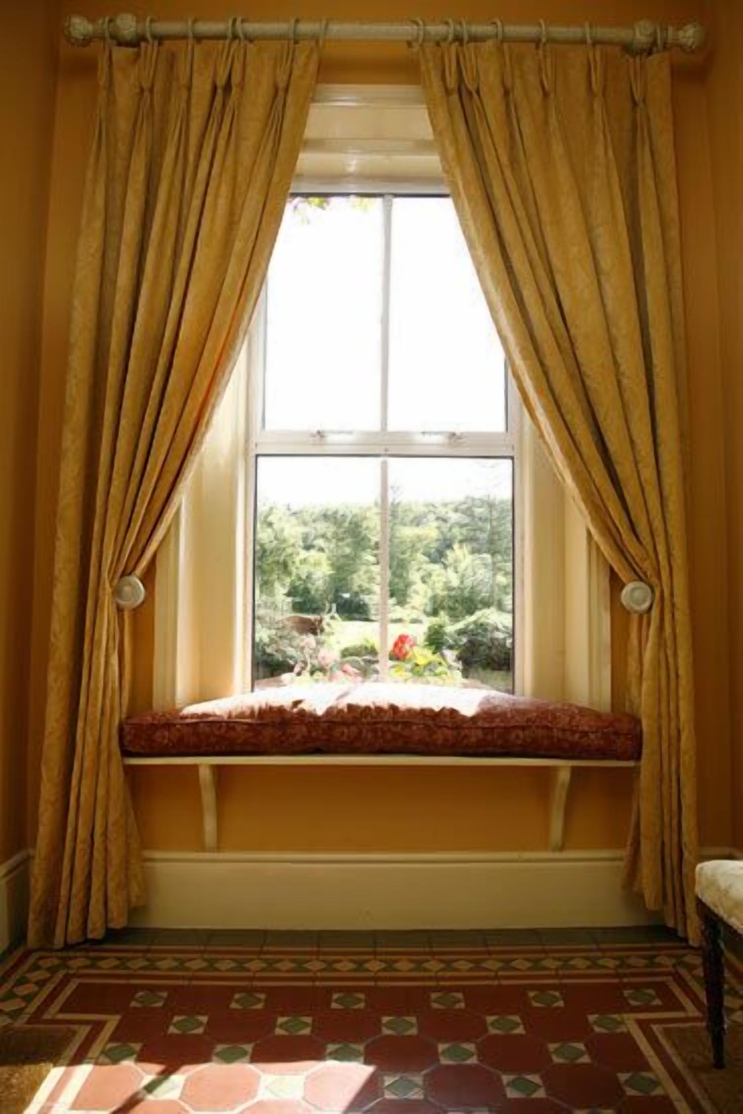 A room with a window, curtains, a bench, and a patterned floor.