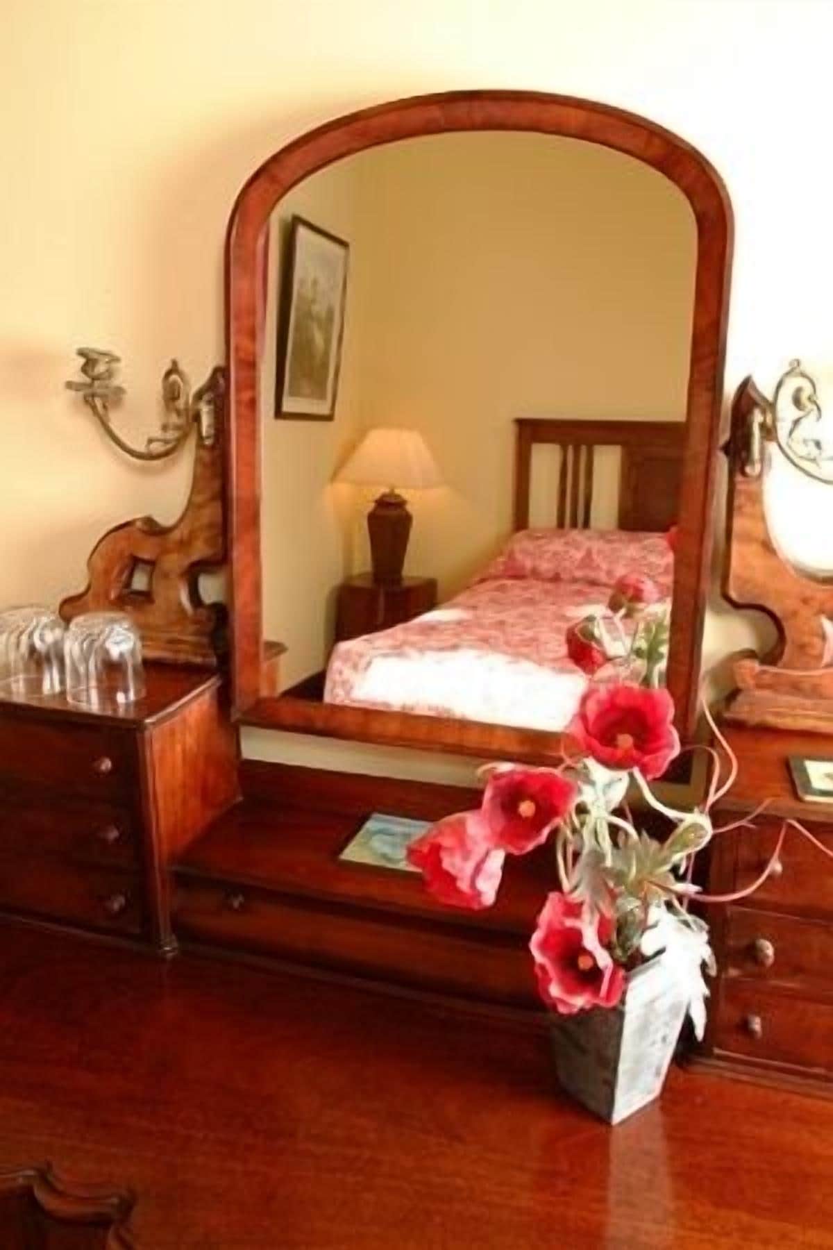 A bedroom with a wooden bed, a mirror, a nightstand, a lamp, and a vase with flowers.