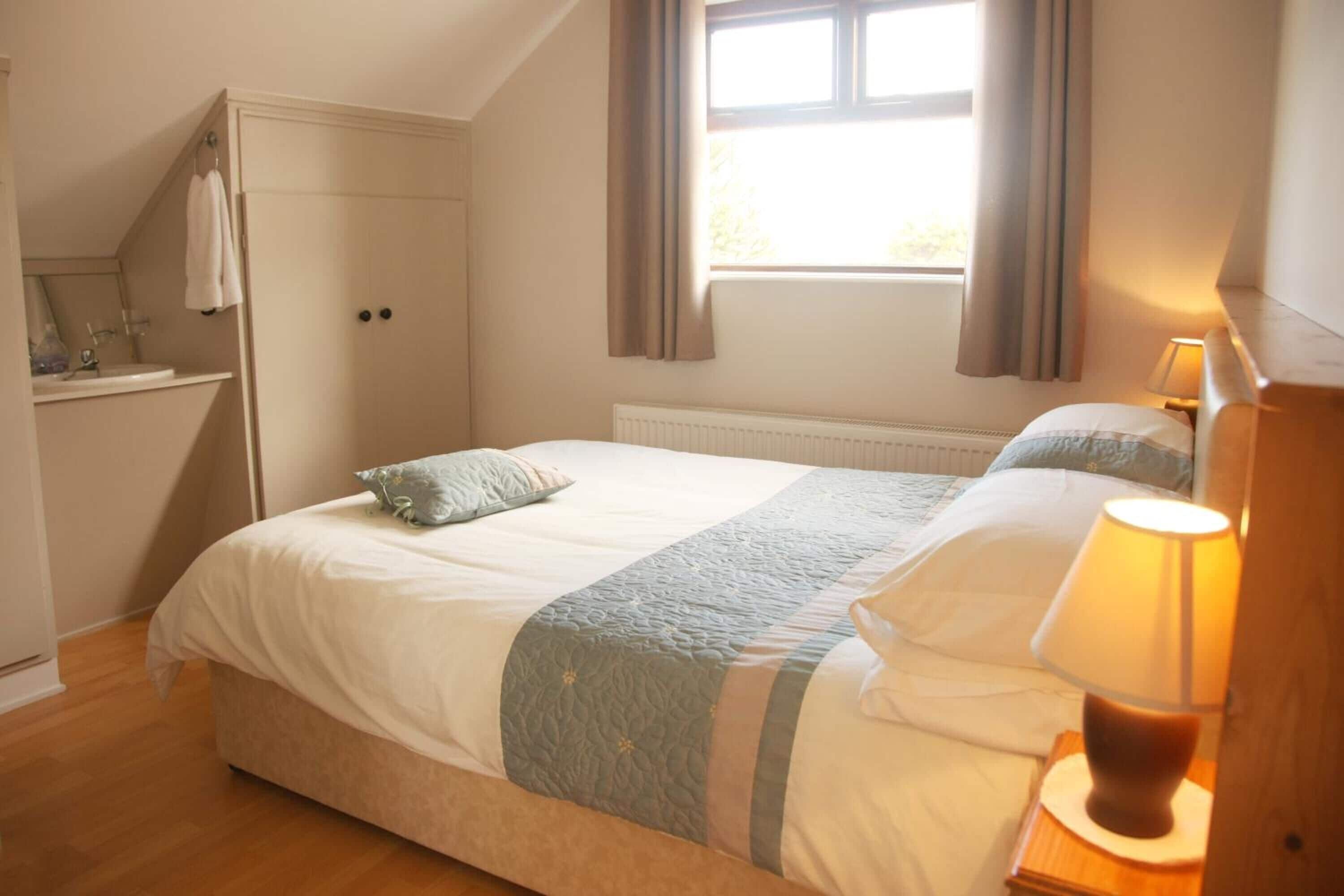 A bedroom with a bed, a nightstand with a lamp, a window with curtains, and a built-in wardrobe.