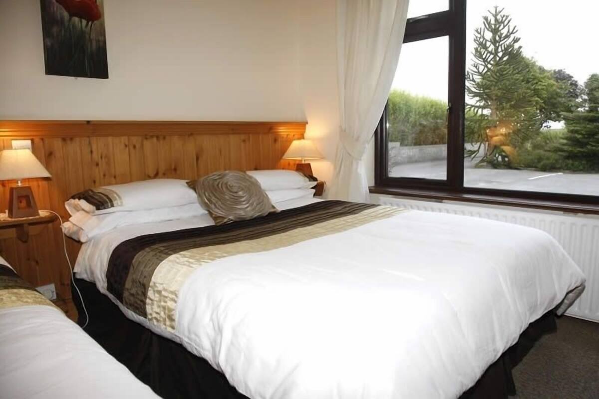 Quarto de hotel com cabeceira de madeira, duas mesas de cabeceira com luminárias, uma cama grande com roupa de cama branca e uma janela com vista para árvores.