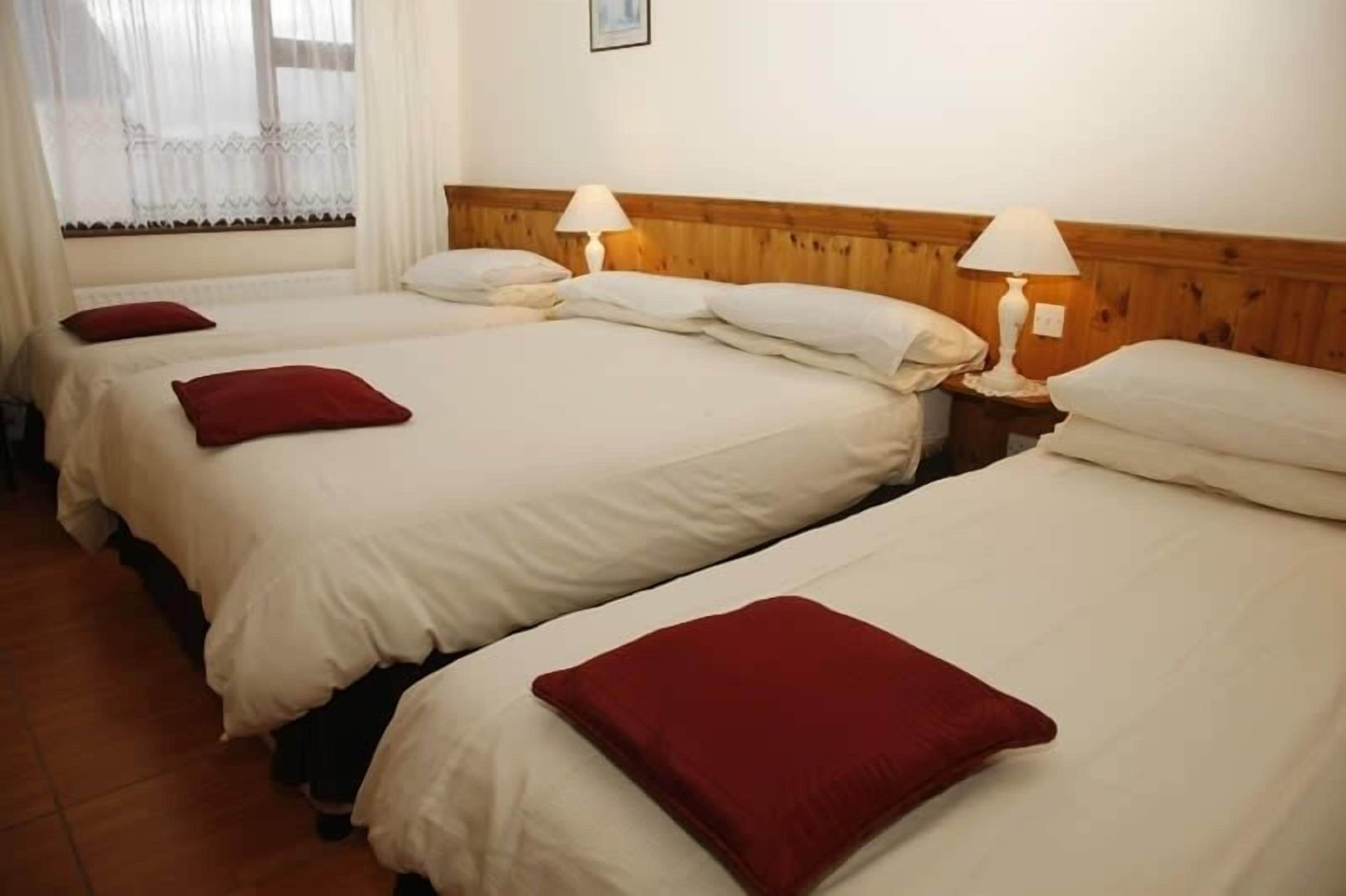 Habitación de hotel con tres camas individuales, cada una con un almohadón rojo y cabecero de madera. Dos lámparas de noche en cada mesita.