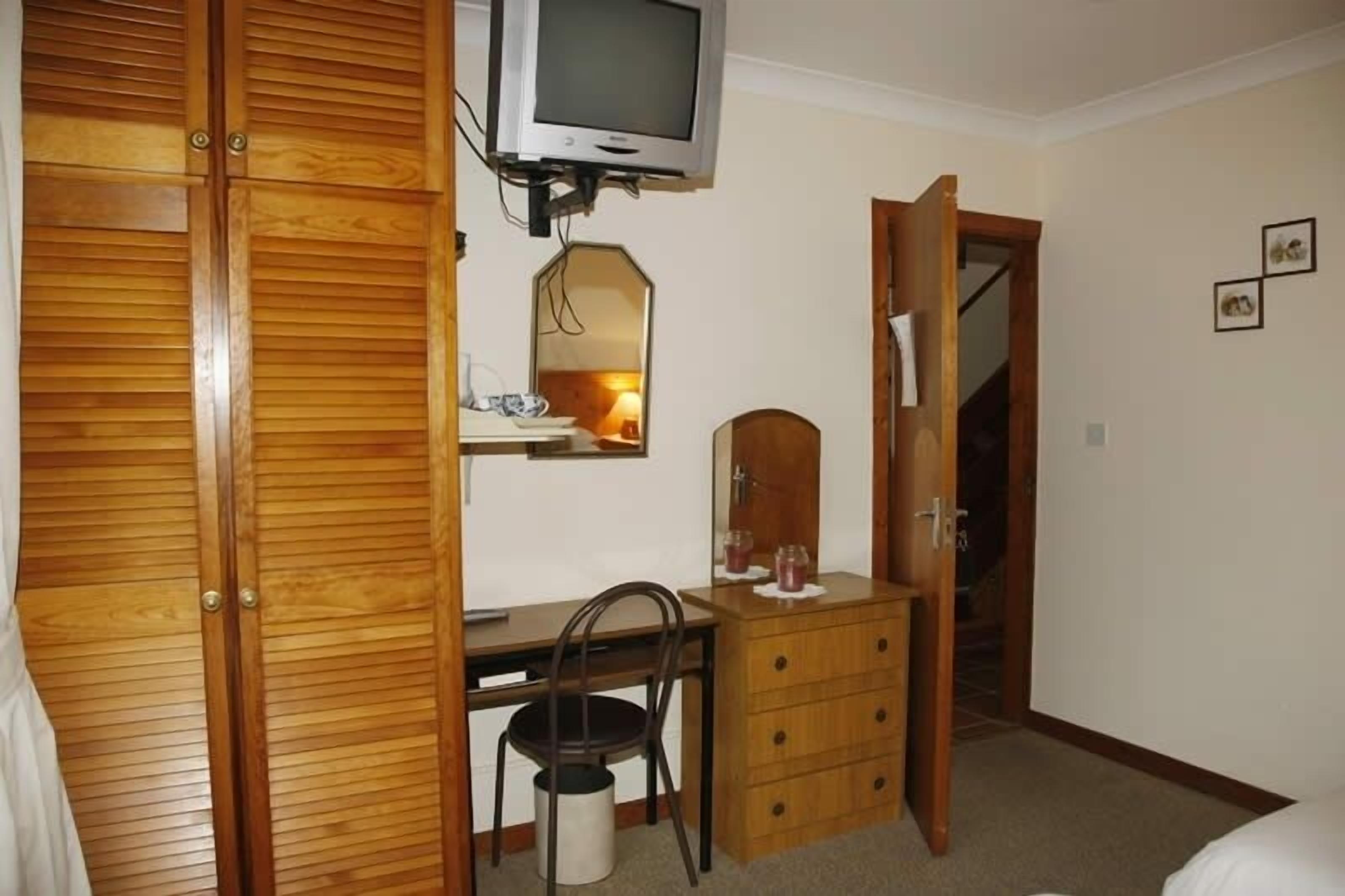 Ein Schlafzimmer mit einem Holzkasten, einem Schreibtisch mit Stuhl, einer Kommode, einem an der Wand befestigten Fernseher und einem Spiegel über der Kommode.