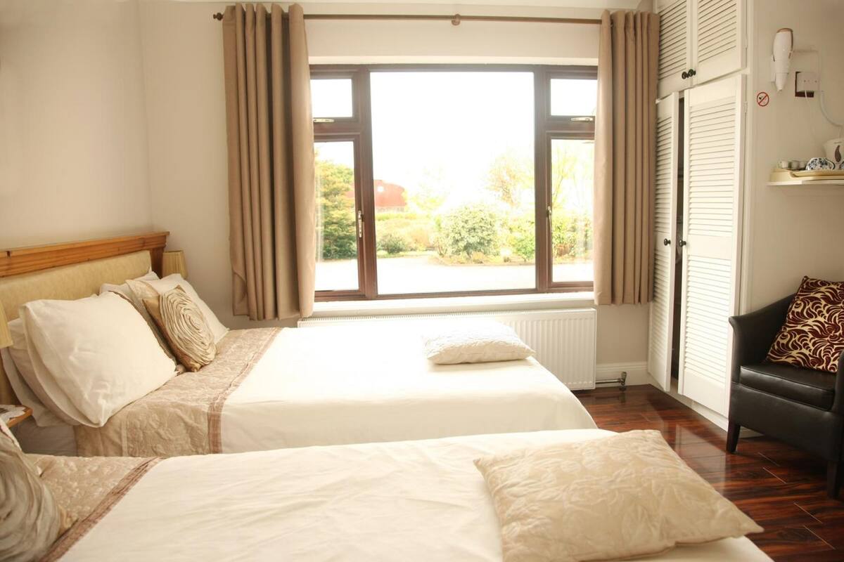 Quarto de hotel com duas camas, janela com cortinas, radiador e uma cadeira preta com almofada estampada.
