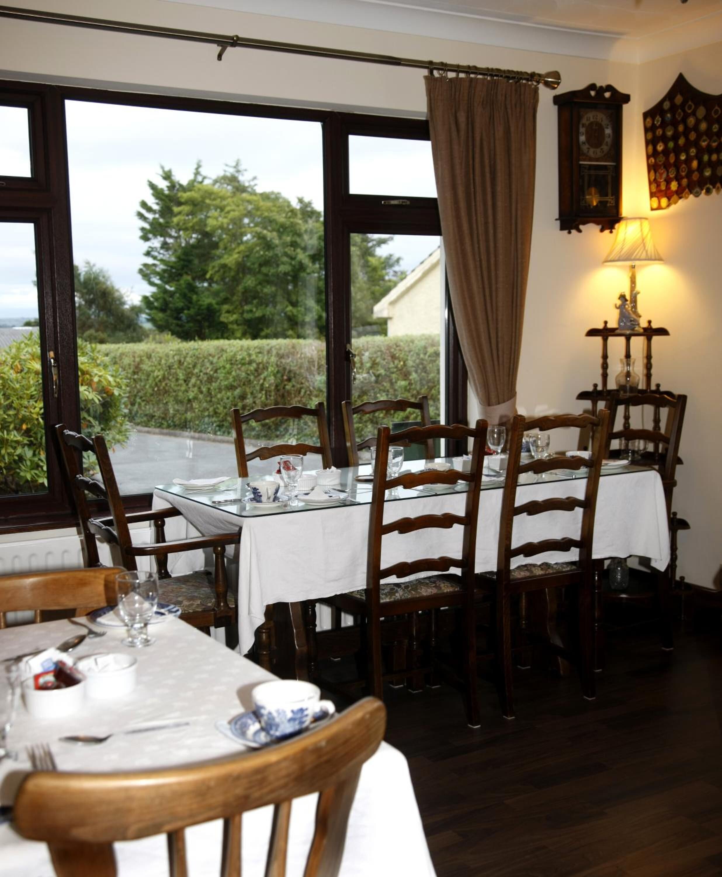 Un coin repas avec une table dressée pour quatre personnes, des chaises en bois, une fenêtre avec des rideaux et une horloge fixée au mur.