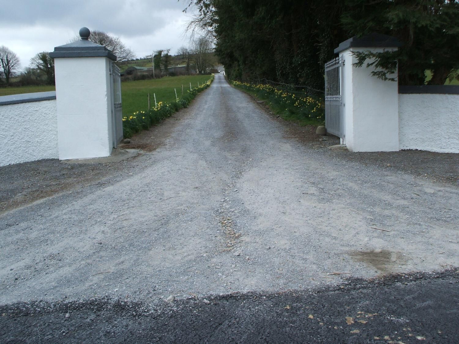 Une allée de gravier mène à une entrée fermée par un portail, avec des murs blancs et un portail en métal.
