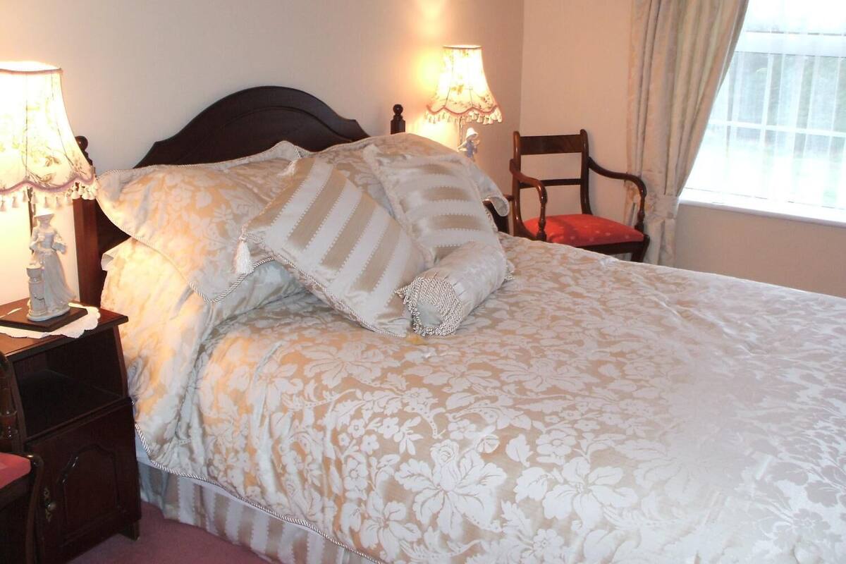Un lit bien fait, avec une literie à motifs floraux, deux lampes de chevet et une chaise agrémentée d’un coussin rouge.