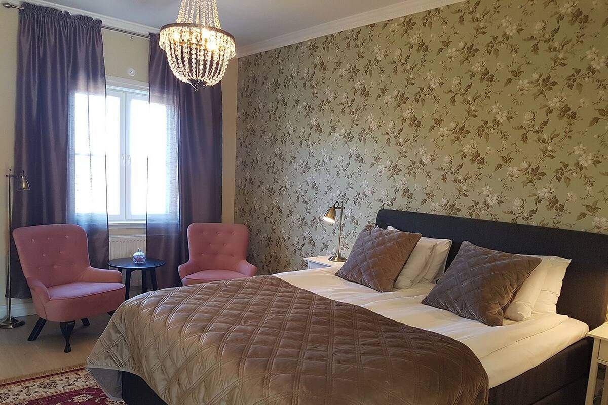 A bedroom with a large bed, two pink armchairs, a chandelier, and floral wallpaper.