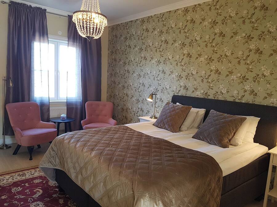 A bedroom with a large bed, two pink armchairs, a chandelier, and floral wallpaper.